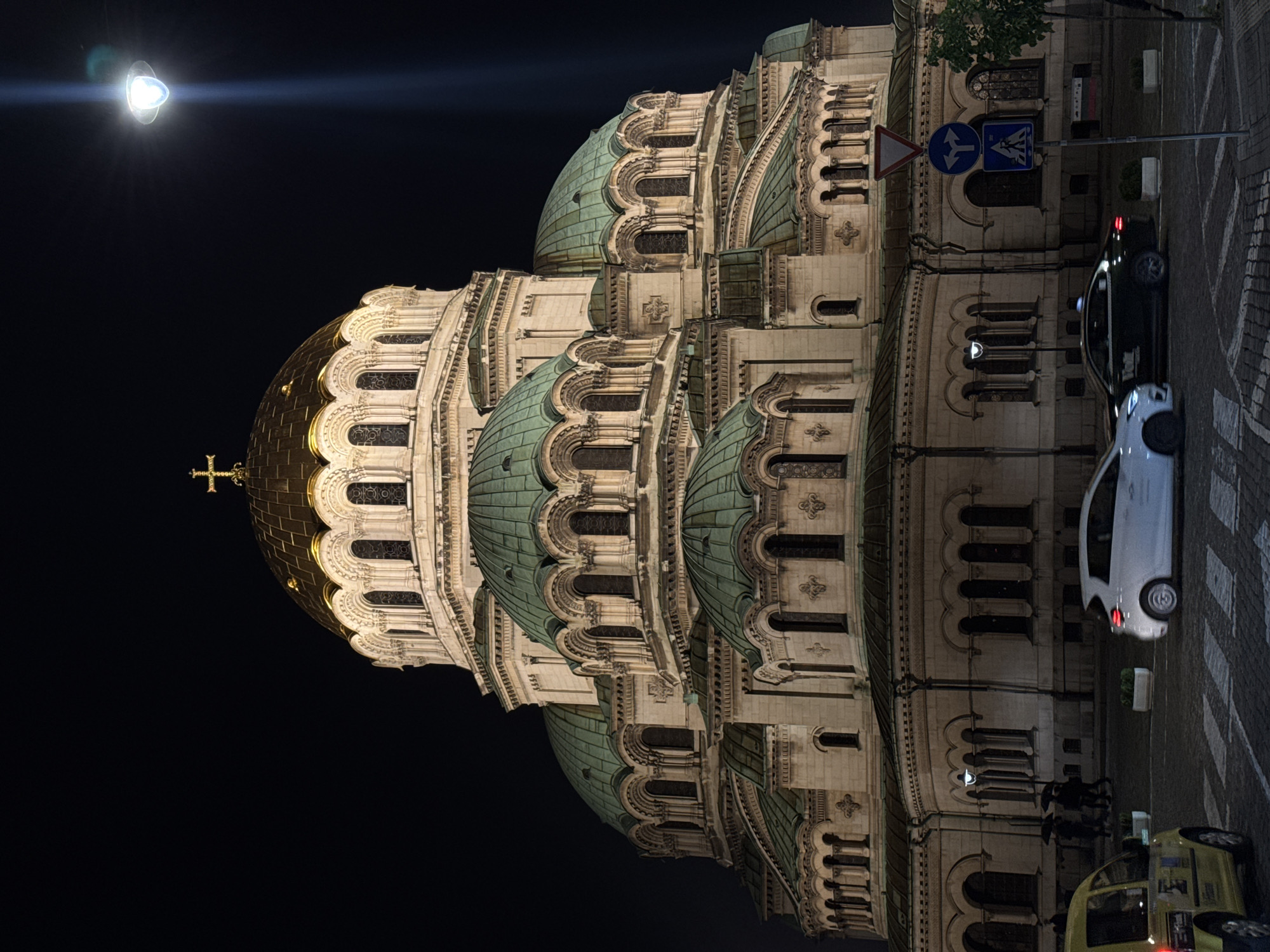 At night, a low-angle, rotated view showcases the Alexander Nevsky Cathedral in Sofia, Bulgaria. The cathedral's golden dome is prominent, brightly lit against the dark sky, and topped with a cross. Several tiered, green-domed structures descend from the main dome. The facade of the cathedral, rendered in light-colored stone, is ornamented with archways, windows, and decorative carvings.
Adjacent to the cathedral, on what appears to be a pedestrian walkway, two cars are parked: a white one and a black one. Further down, a yellow car is parked. A street sign is visible, indicating pedestrian crossing. There are two people walking on the pedestrian walkway.
A streetlight is prominently positioned on the left side of the frame. The overall lighting suggests a nighttime setting.