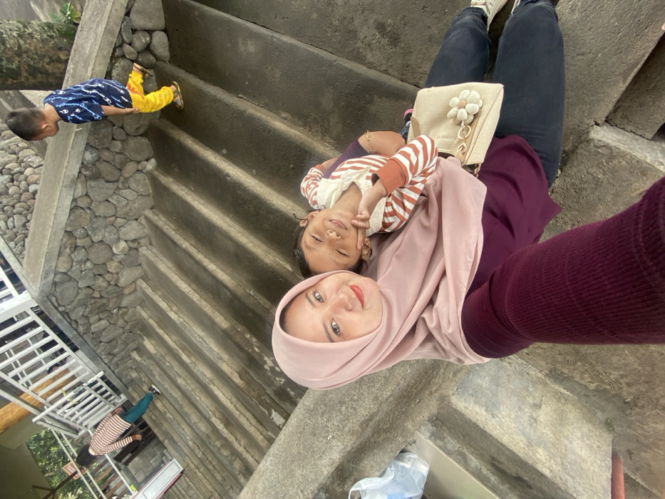 A selfie of a woman wearing a pink hijab and a burgundy top. A child rests on her chest, smiling. They are on concrete steps. A small purse sits on her lap. Another child in a blue shirt and yellow pants is on an upper part of the steps. A partial view of white railings and a building is visible in the background.