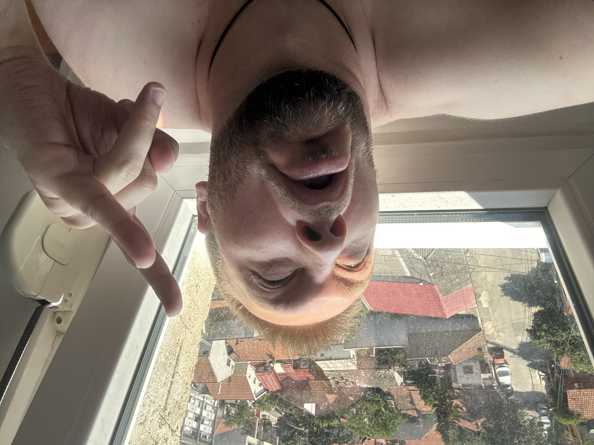 This is a selfie taken from an unusual perspective, likely by a person looking up at the camera through a window. The subject is a man with a beard and fair hair. He is positioned upside down in the frame, with his face close to the camera. His mouth is open, and his eyes are partially closed, giving him a somewhat surprised or playful expression.
A hand with clearly defined fingers and fingernails is visible in the upper left portion of the image, with the index finger and thumb extended, possibly gesturing. The subject appears to be wearing a dark cord around his neck.
The background, seen through a dirty window, shows a view of rooftops of buildings, suggesting an urban setting. The architecture appears to be of European style, with red-tiled roofs common. Trees and a street with parked cars are also visible, indicating it's daytime. The lighting suggests it's a sunny day. The overall impression is a candid and informal moment.