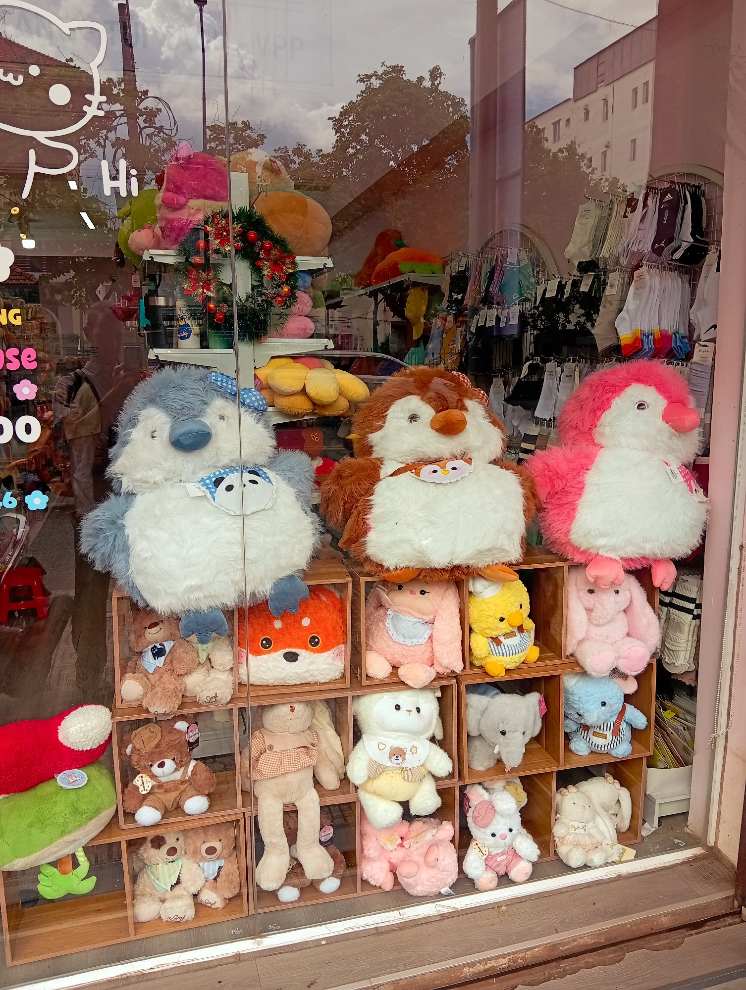 A shop window in Da Lat, Vietnam, displays a collection of plush toys arranged in wooden cubbies. Prominently featured are three large bird-like plushies in shades of blue, brown, and pink. Below them, smaller stuffed animals like bears, rabbits, and an elephant occupy the cubbies. Reflective glass captures the city's exterior, mirroring the surroundings. The store appears to sell hosiery.