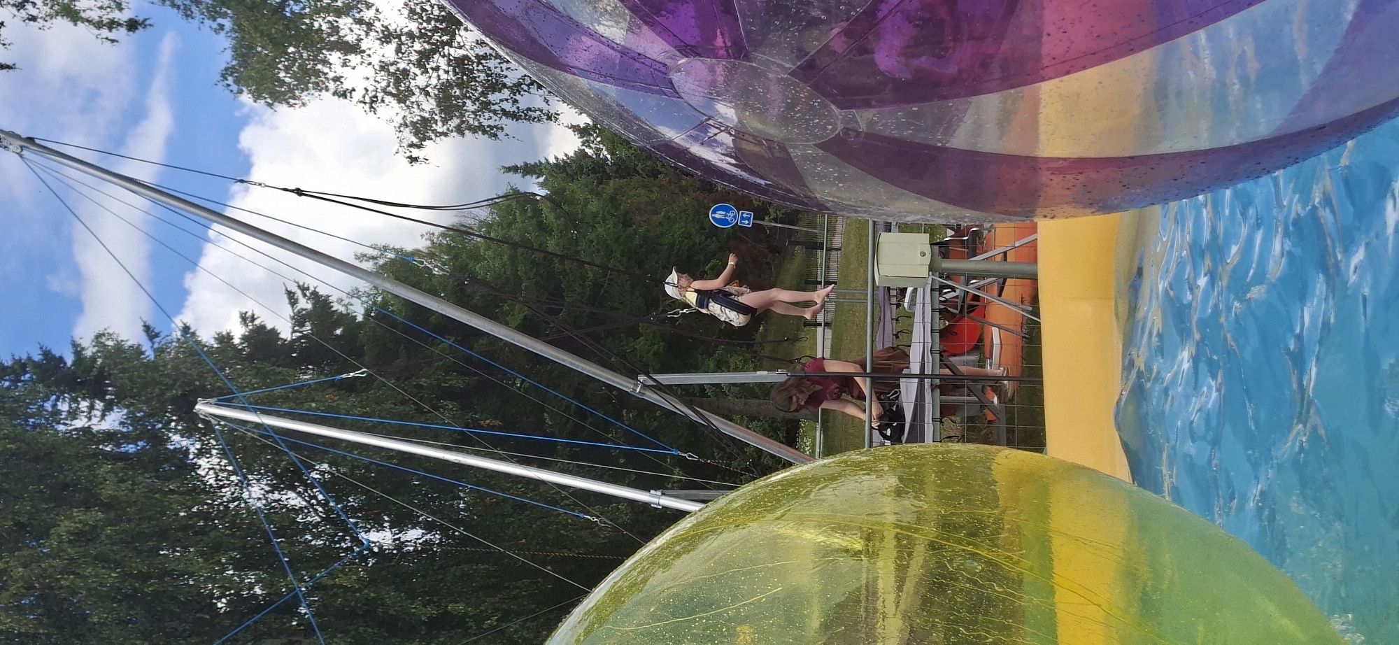 The image shows an outdoor recreational activity involving two large inflatable balls, one yellow and one purple, floating in blue water. In the background, a bungee trampoline setup is visible. A young girl in a harness is suspended mid-air by bungee cords, wearing a white cap and a patterned outfit. Below her, another person, likely an attendant, is seated at a table and appears to be adjusting something, wearing a maroon shirt and brown skirt. The scene is set against a backdrop of green trees and a cloudy blue sky. A sign with pictograms indicating pedestrian and bicycle paths is visible behind the suspended girl. The overall setting suggests a park or a summer activity center. The image is captured from a low angle, partially obscuring the foreground by the large inflatable balls.