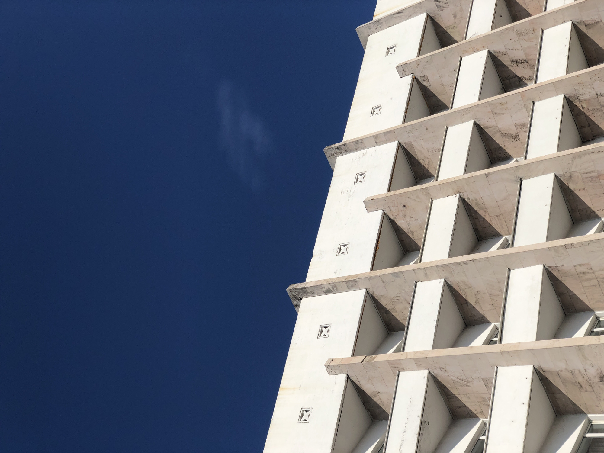 An upward shot captures a segment of a white, vertically tiered building against a deep blue sky, likely in Krasnoyarsk, Russia. The architectural style features projecting tiers, creating angular shadows and a repeating pattern of white and dark triangular shapes. Each level has decorative square recessed elements. A wisp of cloud is visible in the sky. The building’s surface appears textured, possibly due to the material or aging. The composition emphasizes geometric shapes and the contrast between the building and the sky.