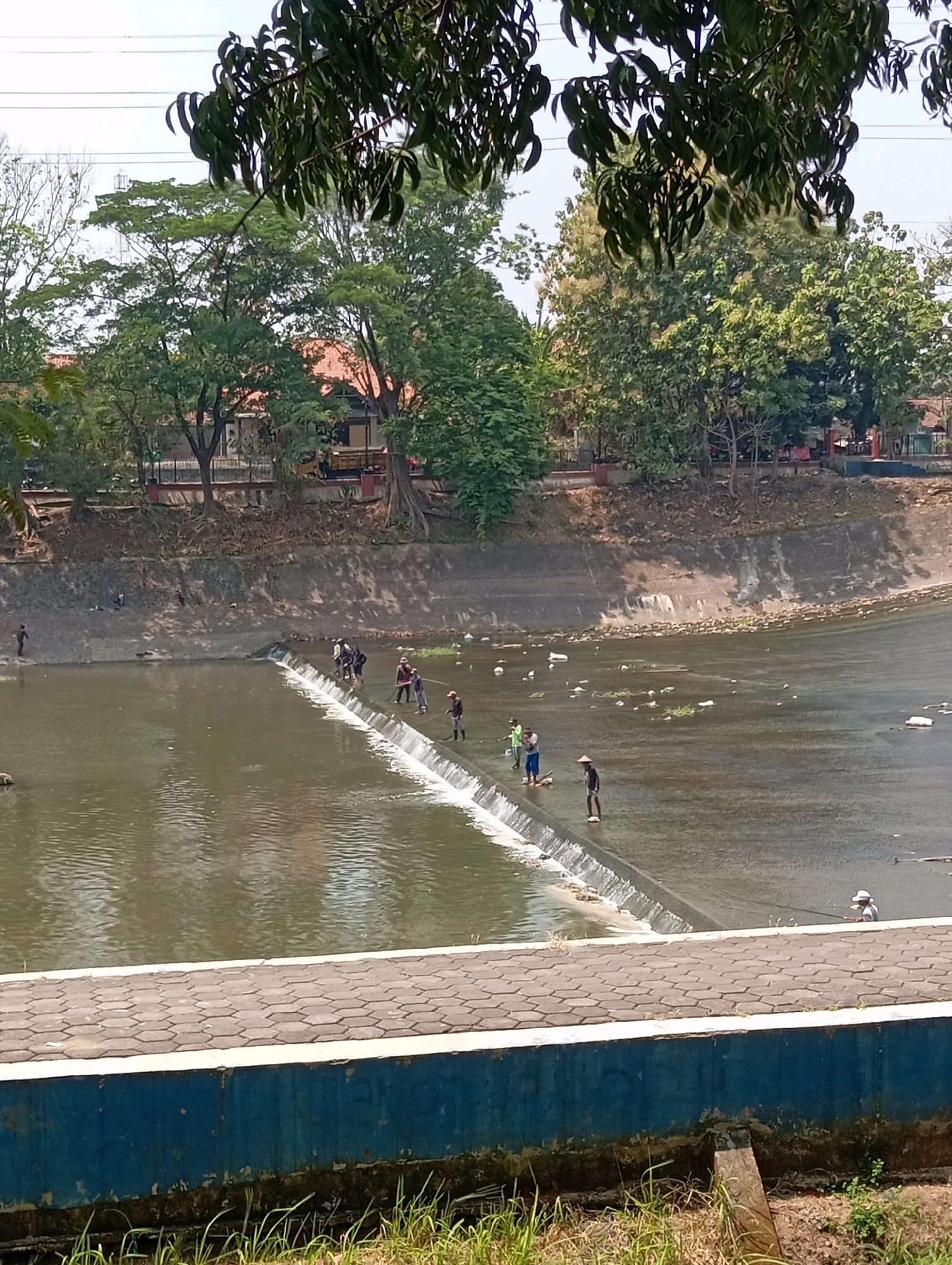 The image depicts a concrete weir in a river, likely part of a water management system in Semarang, Indonesia. Several individuals are present on the weir, engaged in fishing. The water is murky and contains some debris, suggesting it may be a polluted urban waterway. Lush trees and buildings line the riverbank in the background, indicating an inhabited area. The foreground shows a paved walkway and a blue concrete wall, likely part of a public access area or embankment. The overall scene suggests a common urban landscape where recreation and infrastructure coexist.
