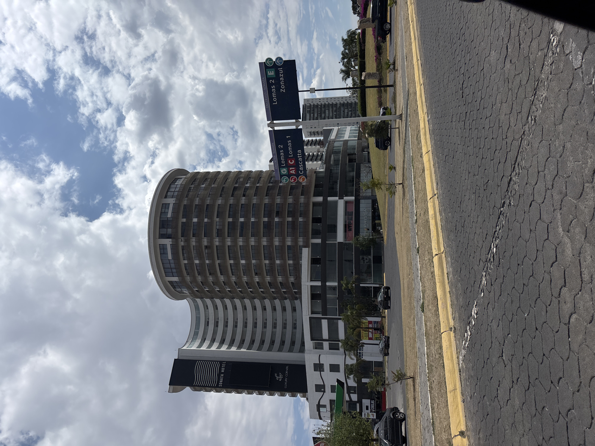 The image shows a street scene in San Bernardino Tlaxcalancingo, Mexico. Prominent features include a tall, curved building identified as "Sonata Hotel" and signage indicating "Lomas 2" and "Lomas 1." A street sign also points towards "Zonaazul." The street is paved with a textured surface. Several vehicles are parked along the side of the road. The sky is partially cloudy.