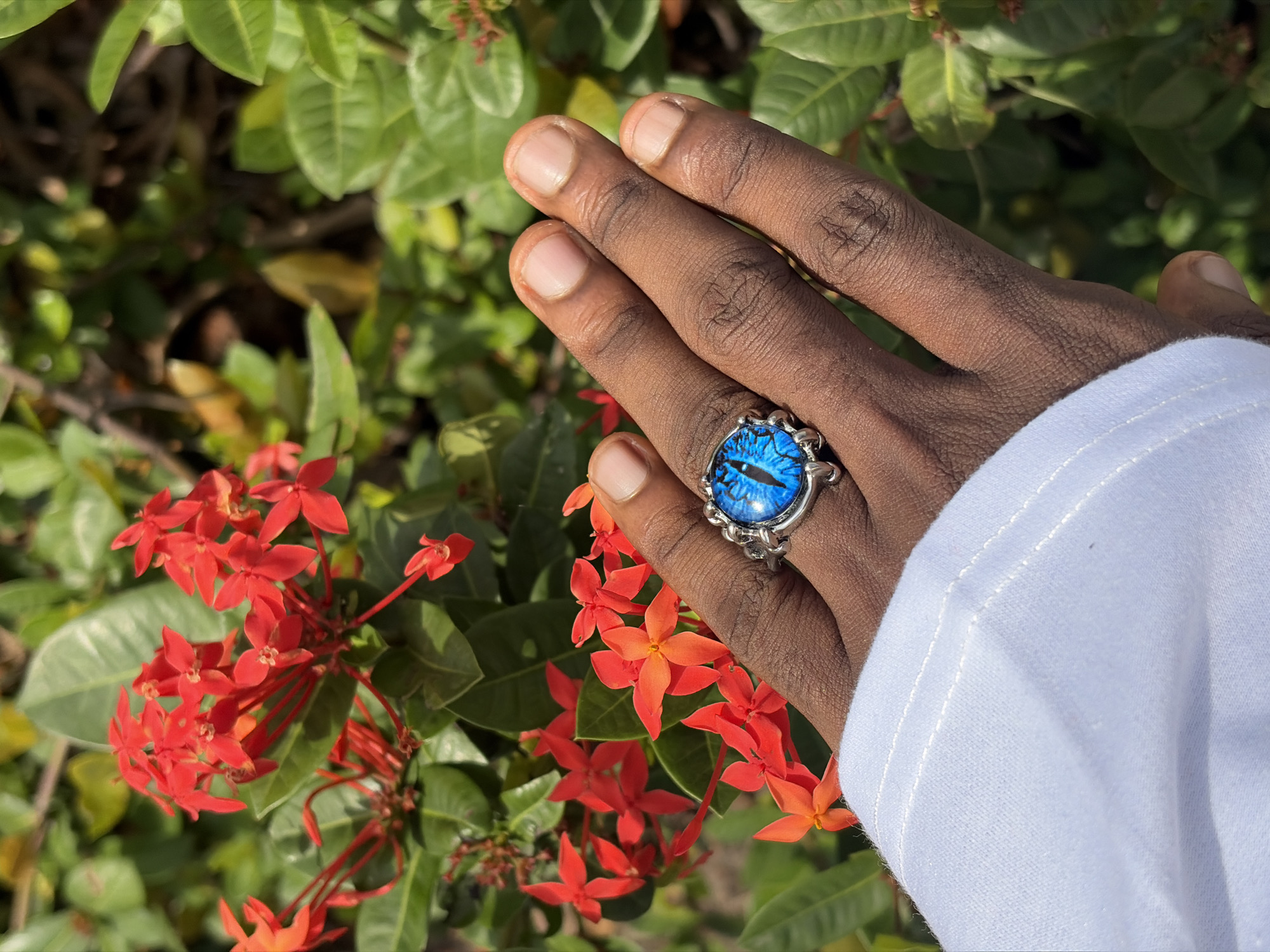 A close-up shot focuses on a person's hand adorned with a striking silver ring featuring a blue dragon eye design. The hand, with dark skin, is positioned against a backdrop of lush green foliage and vibrant red ixora flowers. The person is wearing a white garment, likely a shirt, which covers part of the arm. The image is captured outdoors, suggesting a natural setting. The presence of red ixora flowers and green leaves indicates a tropical or subtropical climate. The specific location of Igboefon, Nigeria, is mentioned as the setting for this image.