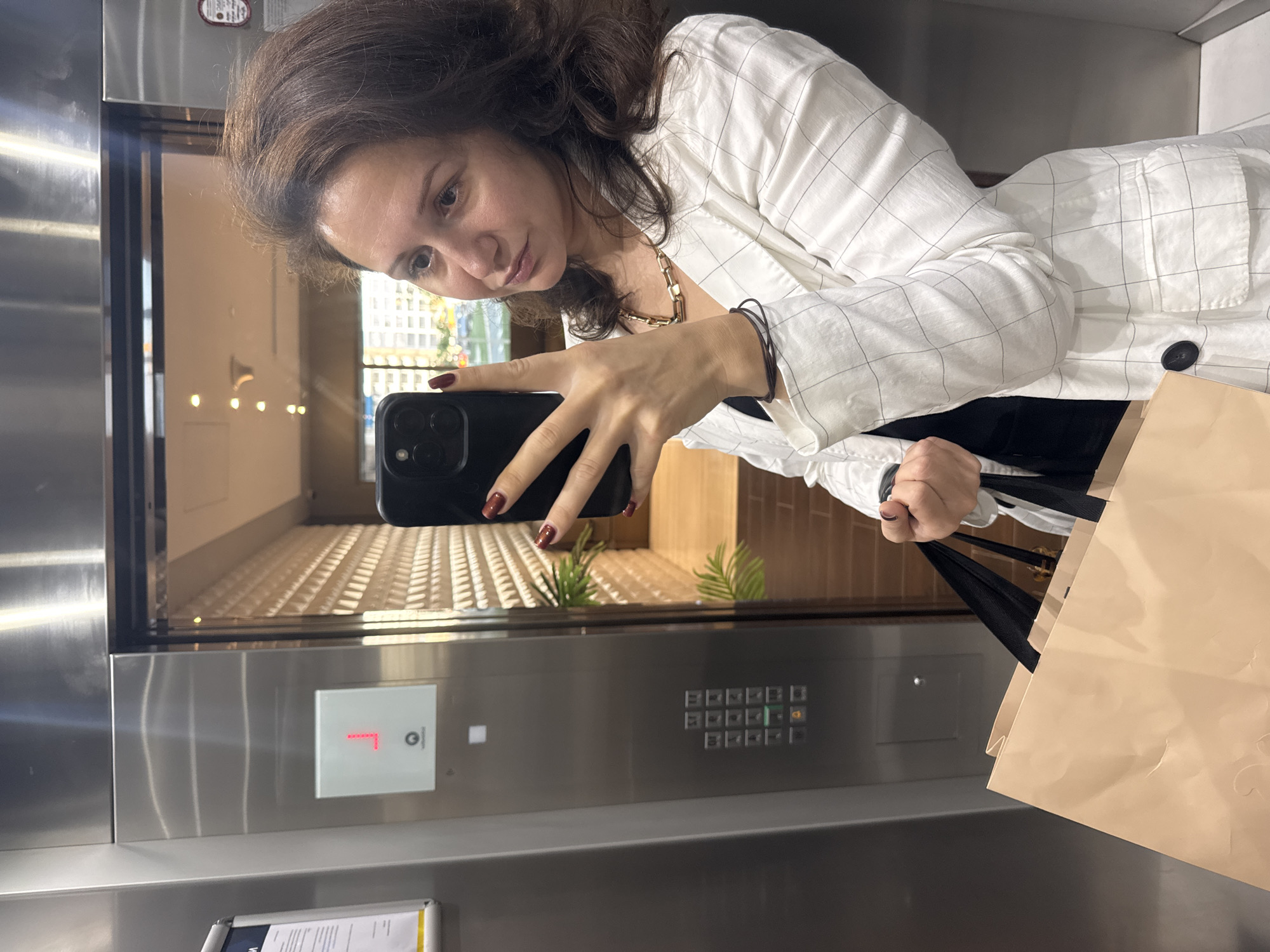 A woman is taking a selfie in an elevator. She is wearing a white, checkered jacket and a gold necklace. She is holding a black phone in her left hand, and a brown shopping bag in her right hand. The elevator is paneled with metal, and the buttons show that the elevator is on the first floor. A reflection in the elevator door shows a modern lobby with plants and a view of a city street outside, which is likely in Dubai, United Arab Emirates, based on the architectural style and landscaping visible in the reflection.