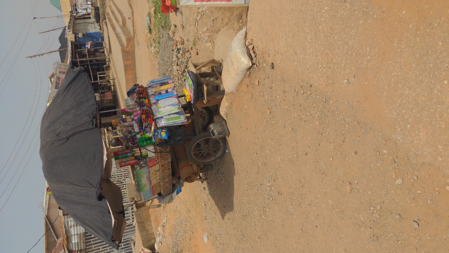 The image, taken in Ado, Nigeria, depicts an outdoor street market scene on an unpaved, dirt and gravel road under a clear, sunny sky.
In the foreground, a mobile market stall or cart is prominently featured, sheltered by a large, dark grey umbrella. The stall is laden with a diverse array of goods, including stationery items such as rulers, pens, and pencil cases, along with various toiletries like shampoo bottles, and other small household products. A white sack rests on the ground beside the cart.
In the background, a row of informal shops or structures line the street, some constructed with corrugated metal roofs. Utility poles and wires are visible against the bright sky. A sign above one of these background shops clearly reads "MUMMY SHOP." The lighting suggests it is daytime, possibly late morning or early afternoon, with shadows cast by the cart and umbrella. The scene portrays a typical bustling, informal commercial area. No specific human activity is visibly captured, though the setup clearly indicates vending.