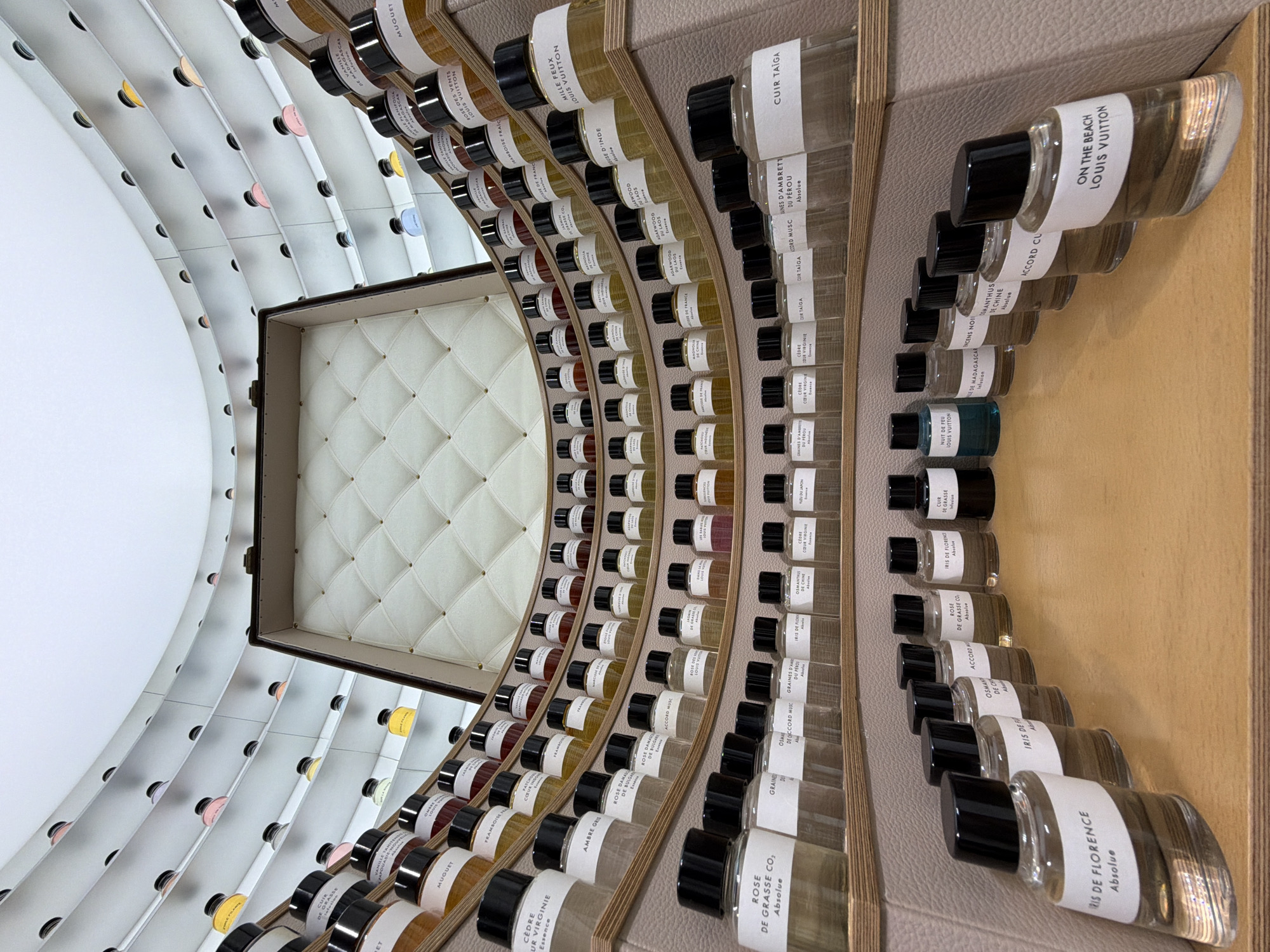 This indoor shot depicts a curated perfume display, likely located in a retail store or museum. The perfumes, predominantly by Louis Vuitton, are arranged in a concentric, curved pattern around a central case lined with white quilted material. The bottles feature black caps and white labels with black text. The display shelves are a light wood tone, set against walls with pale beige or gray surfaces. The display includes labels such as "CUIR TAIGA," "ON THE BEACH LOUIS VUITTON," and "IRIS DE FLORENCE." The composition includes many small, transparent perfume bottles, some filled with colored liquids. The labels suggest the origin of some ingredients like "Yuzu du Japon," "Boisse de France," "Agarwood du Laos," and the country "China." The arrangement suggests a deliberate attempt to showcase a wide range of scents in a visually compelling manner. It can be inferred from the OCR text that this display is likely a Louis Vuitton perfume collection, and the labels suggesting places of origin indicate that it might be related to a travel-themed collection. The specific location within the Jing'an District, China, cannot be precisely determined from the visual data provided.