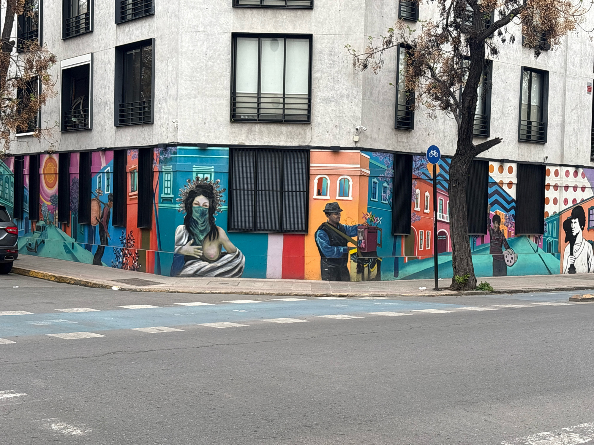 This is a medium-shot, eye-level, full-color photograph taken outdoors on an overcast day in the city of Santiago, Chile. The image captures a corner of a modern building with a vibrant mural covering its lower facade. The building has a light gray, concrete exterior with several dark-framed windows. A leafless tree with dried foliage stands in front of the building, partially obscuring the mural and the upper floors of the structure.
The mural is colorful and eclectic, featuring various figures and scenes. On the left, a woman with dark hair and a green mask is depicted with her bare breasts visible. To her right, a man wearing a hat and a dark vest over a blue shirt is shown holding a tool. Further to the right, a figure with dark hair and a white shirt is seen holding a guitar. The mural also includes stylized buildings, geometric patterns, and a bicycle symbol on a blue signpost.
In the foreground, a street corner with a crosswalk is visible. The crosswalk is marked with blue paint and white squares. A small, white piece of trash is on the sidewalk near the mural. A dark-colored car is parked on the left side of the image, partially visible. The overall environment suggests an urban setting with an artistic touch.