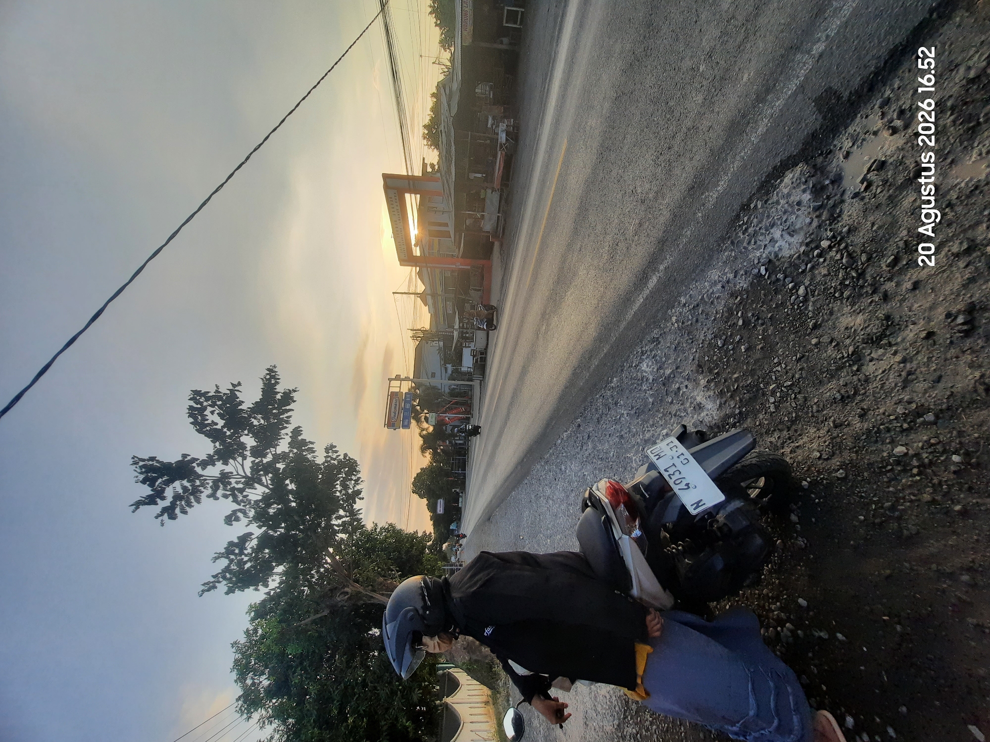 A person wearing a black jacket, blue pants, and a black helmet sits on a scooter parked on the side of a road. The scooter has a license plate that reads "N 4931 MO 01-21". The road is paved and appears to be in a semi-urban or rural area. In the background, there are buildings, trees, and utility poles. The sun is setting, casting a warm glow across the sky. A timestamp on the image indicates "20 Augustus 2026 16.52". The location is identified as Sukokerto, Indonesia.