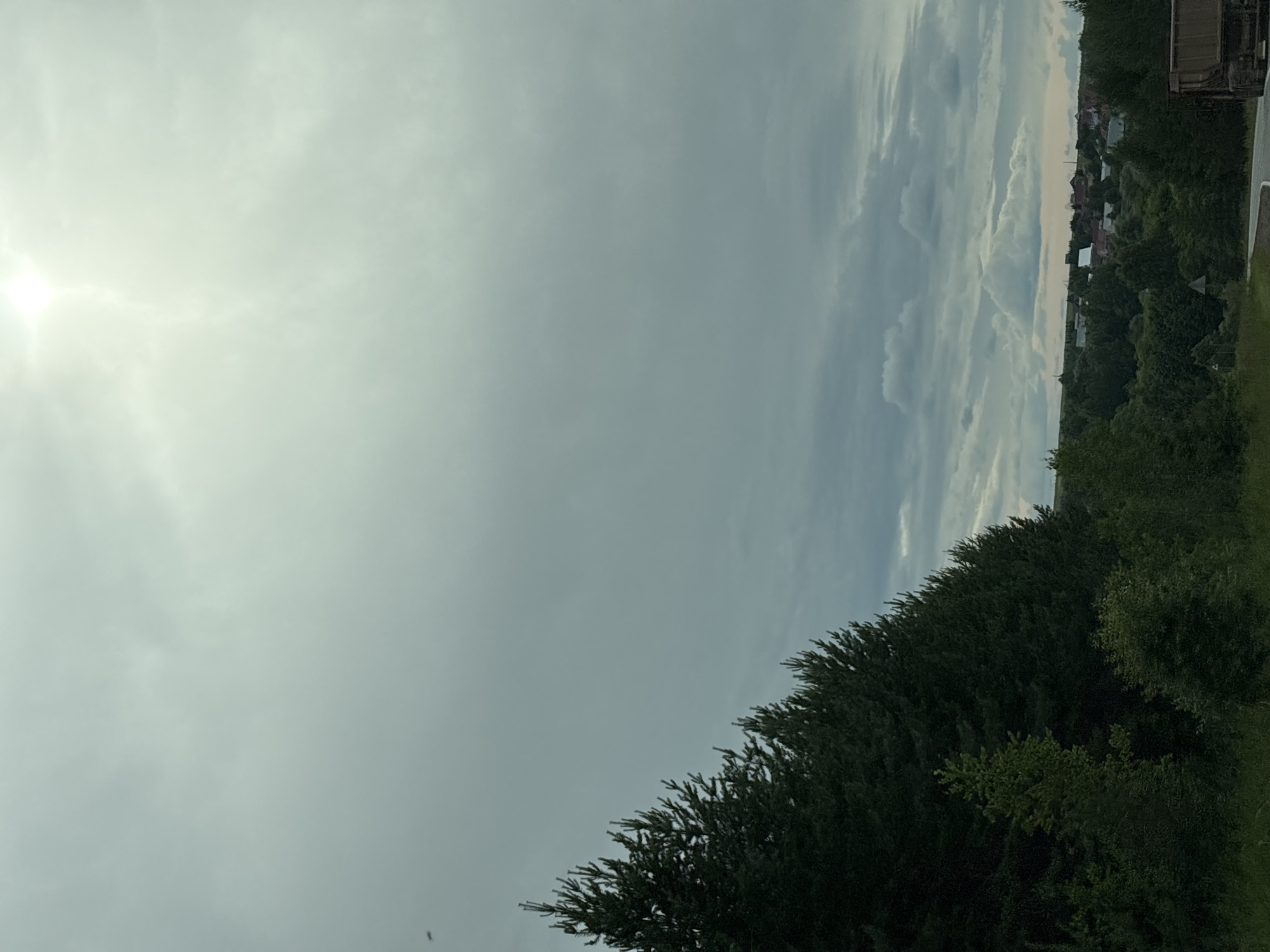 This media file shows a slightly overcast sky with the sun peeking through on the left side. The scene transitions to a view of trees and a distant village with buildings and rooftops. A dark-colored truck is partially visible on the far right edge. The foreground is dominated by lush green trees, appearing dense and well-maintained. The overall impression is a rural or semi-rural landscape under a daytime sky. No people or specific activities are evident, suggesting a still or observational shot. The lighting implies it is either morning or late afternoon, given the angle of the sun. The context provided suggests the location is Забегаловo, Russia. There is no visible text in the image.