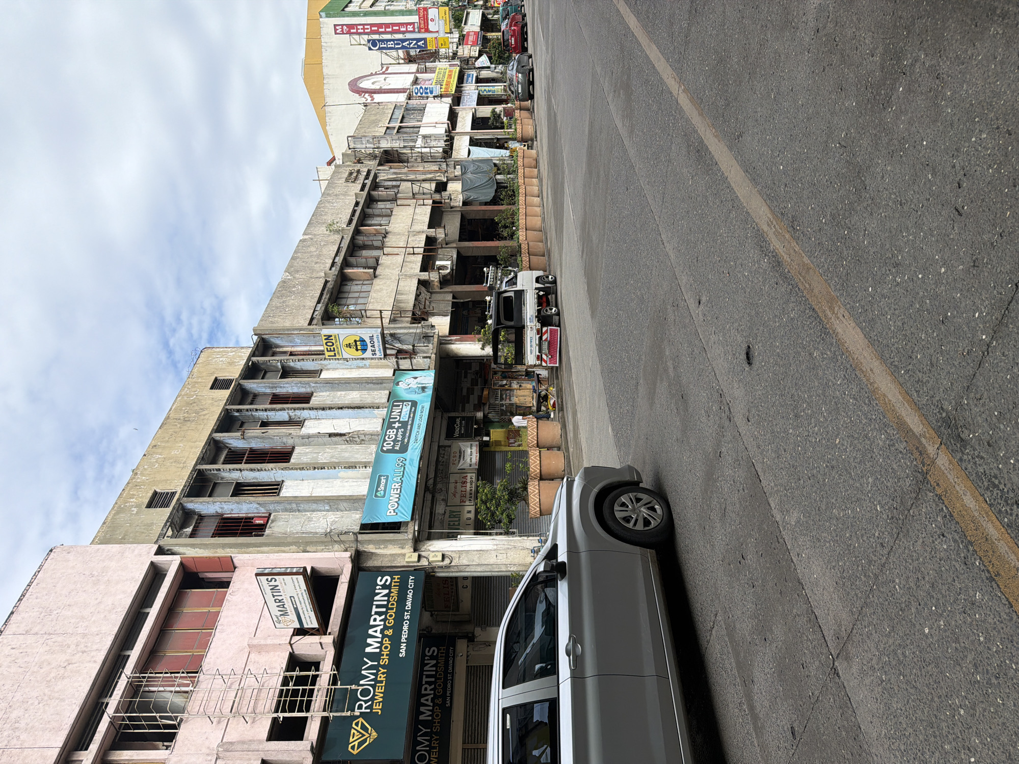 The image captures a street view in Davao City, Philippines, likely taken in the San Pedro Street area. The perspective is slightly angled, showcasing a row of buildings with various commercial establishments on the ground floor.
Prominently visible are the storefronts of "ROMY MARTIN'S JEWELRY SHOP & GOLDSMITH," with signage also bearing the name "MARTIN'S". The buildings appear aged, with visible wear and tear. Additional businesses are visible, including a pawnshop ("Agencia FELISA ION") and signage for cellular services ("Smart POWERALL99").
A silver car is parked on the right side of the street, partially obscuring the view of the storefronts. The road has a faded yellow lane marking. The sky is overcast, casting a diffused light on the scene. Further up the street, other vehicles are visible, along with more commercial signage.