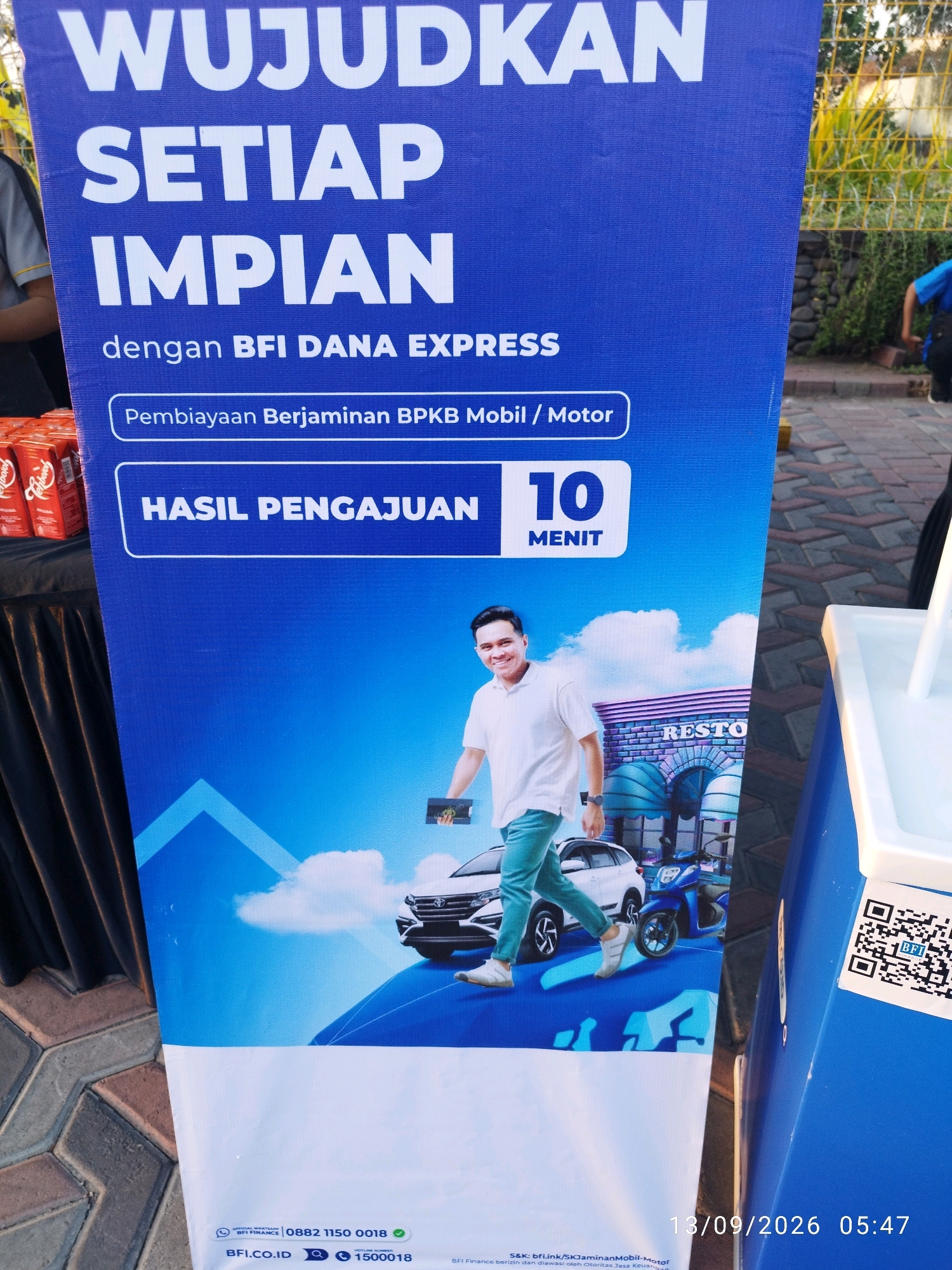 A blue banner with white text is displayed outdoors. The banner reads "WUJUDKAN SETIAP IMPIAN dengan BFI DANA EXPRESS". Below this, it states "Pembiayaan Berjaminan BPKB Mobil / Motor" and "HASIL PENGAJUAN 10 MENIT". A man in a white t-shirt and turquoise pants is depicted walking towards a white car and a blue motorcycle. The background features a stylized building with the word "RESTO" visible. The lower portion of the banner has contact information including a WhatsApp number "0882 1150 0018", a website "BFI.CO.ID", and a hotline number "1500018". A QR code and the date "13/09/2026 05:47" are visible on the right side of the image. The banner is positioned in a paved outdoor area with a slight incline, suggesting it might be in a public space such as a street or park. The scene is likely set in Mojokerto, Indonesia, given the Indonesian language used on the banner.