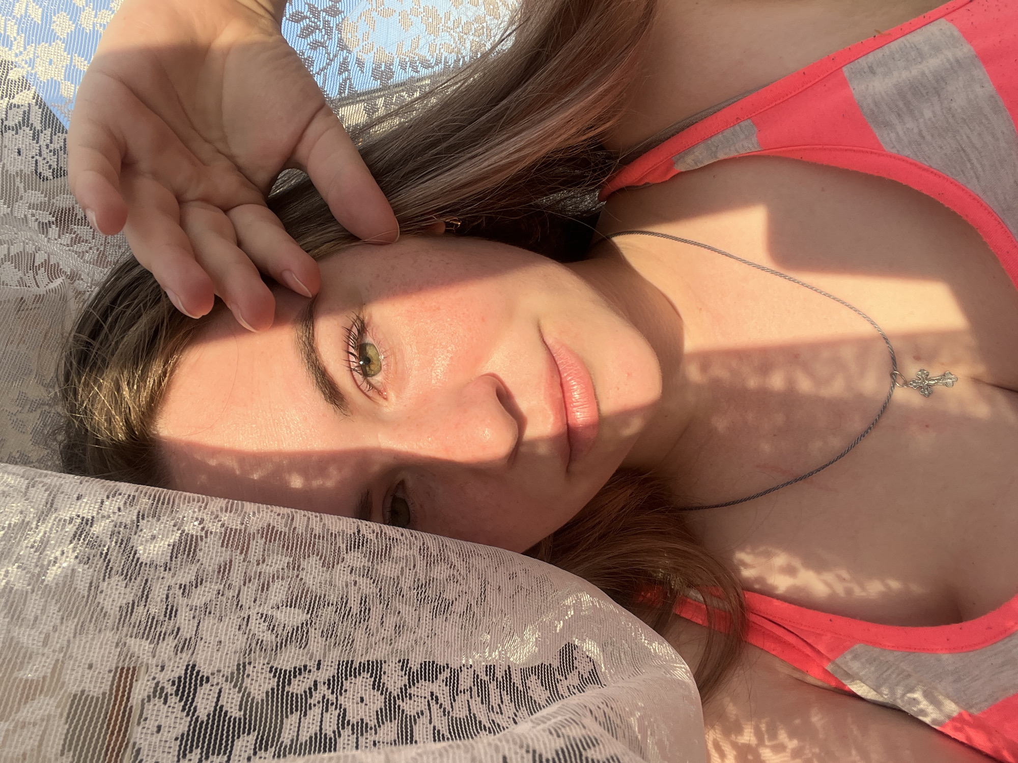 A young woman is lying down on a white lace fabric with light shining on her face and body. She is wearing a pink and gray tank top and a silver necklace with a cross pendant. Her left hand is raised to her forehead. The light creates shadows on her skin and clothes. The location is unknown and there is no evidence of the city of Chaikovsky, Russia.