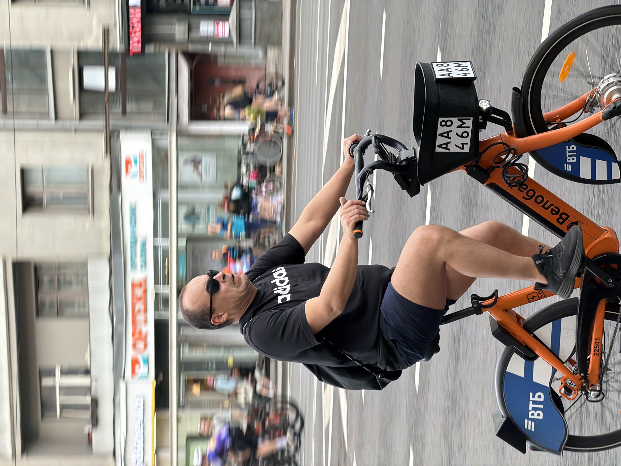 A man is riding an orange and black rental bicycle on a city street. The bicycle is branded "Велобайк" and features "ВТБ" logos, indicating a sponsorship with VTB Bank. The bicycle has a front basket with a license plate "AA8 46M." The man is wearing a black Adidas t-shirt, dark shorts, black sunglasses, and dark shoes. He is riding on a paved street with multiple lanes, possibly a dedicated bike lane. In the background, there are blurred images of other people, buildings, and shop signs, suggestive of a busy urban environment. The location is most likely Moscow, Russia, based on the Велобайк branding.