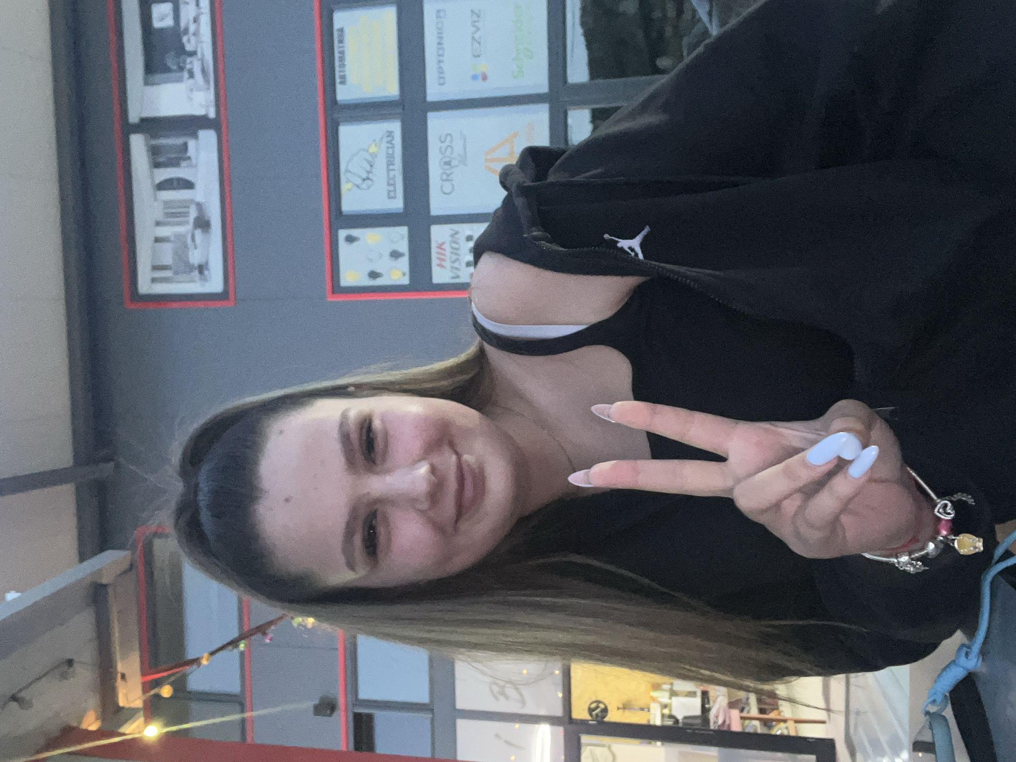 A young woman with light-brown hair pulled back from her face in a ponytail smiles and holds up a peace sign. She wears a black zippered jacket with a white logo on the left side. Her fingernails are painted a light blue. On the wall behind her are several advertisement boards with logos including "AUTOMATIKA", "ELECTRICIAN", "HIK VISION", "OPTONICA", "CROSS", "EZVIZ", and "Schneider." The location appears to be Satovcha, Bulgaria, based on the logos of local businesses. There are storefront windows visible in the background with lights.