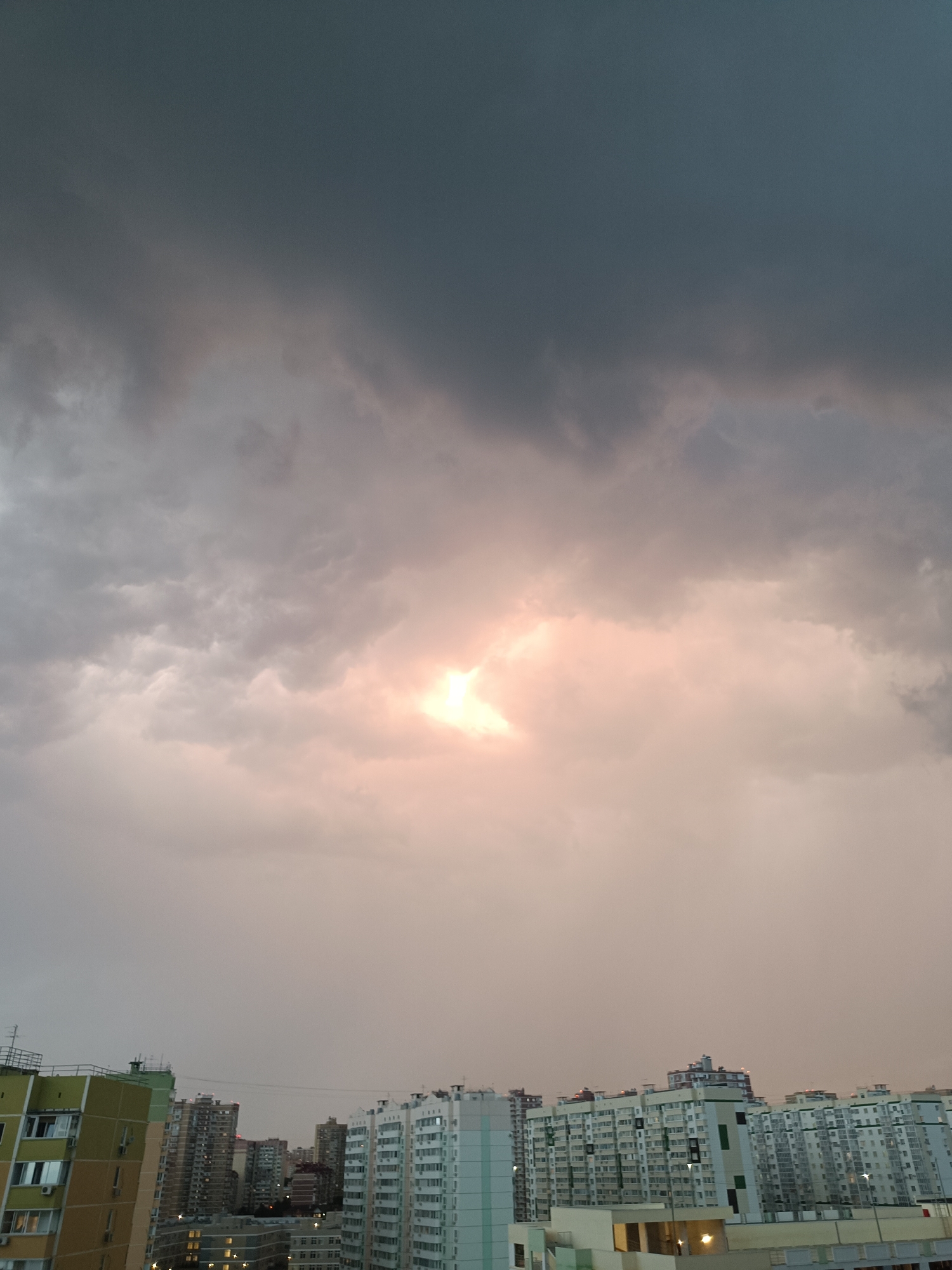 The image captures a dramatic sky over a cityscape. The sky is filled with dark, ominous clouds, with a bright patch of sunlight breaking through the center, casting a warm, pinkish glow. Below the sky, a dense collection of apartment buildings stretches across the horizon. The buildings are of varying heights and architectural styles, with many featuring balconies and visible window units. The overall impression is that of a cloudy day, possibly before or after a storm, with the sun making a brief appearance. The location is identified as the city of Krasnodar, Russia.