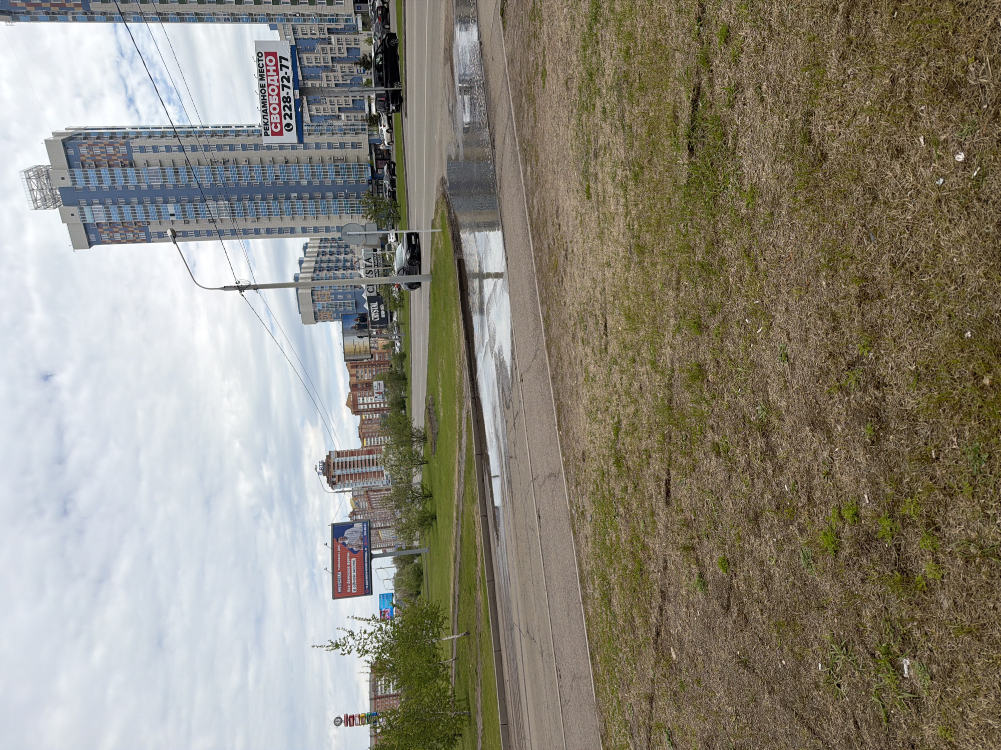 The image depicts a street scene in Krasnoyarsk, Russia, with a view of residential buildings and advertising billboards. The buildings are primarily high-rise structures with blue and white facades. The billboards display Russian text, including advertisements for real estate ("РЕКЛАМНОЕ МЕСТО СВОБОДНО 228-72-77") and medical services ("МЕДИГЕЛФ Работаем с вас все детские врачи в одном месте").
The foreground consists of a road with standing water and a grassy area. The grass appears dry and sparse. The sky is overcast with clouds. There are a few trees and lampposts along the road. Vehicles are parked along the side of the road. The overall environment suggests an urban area after a period of rain.