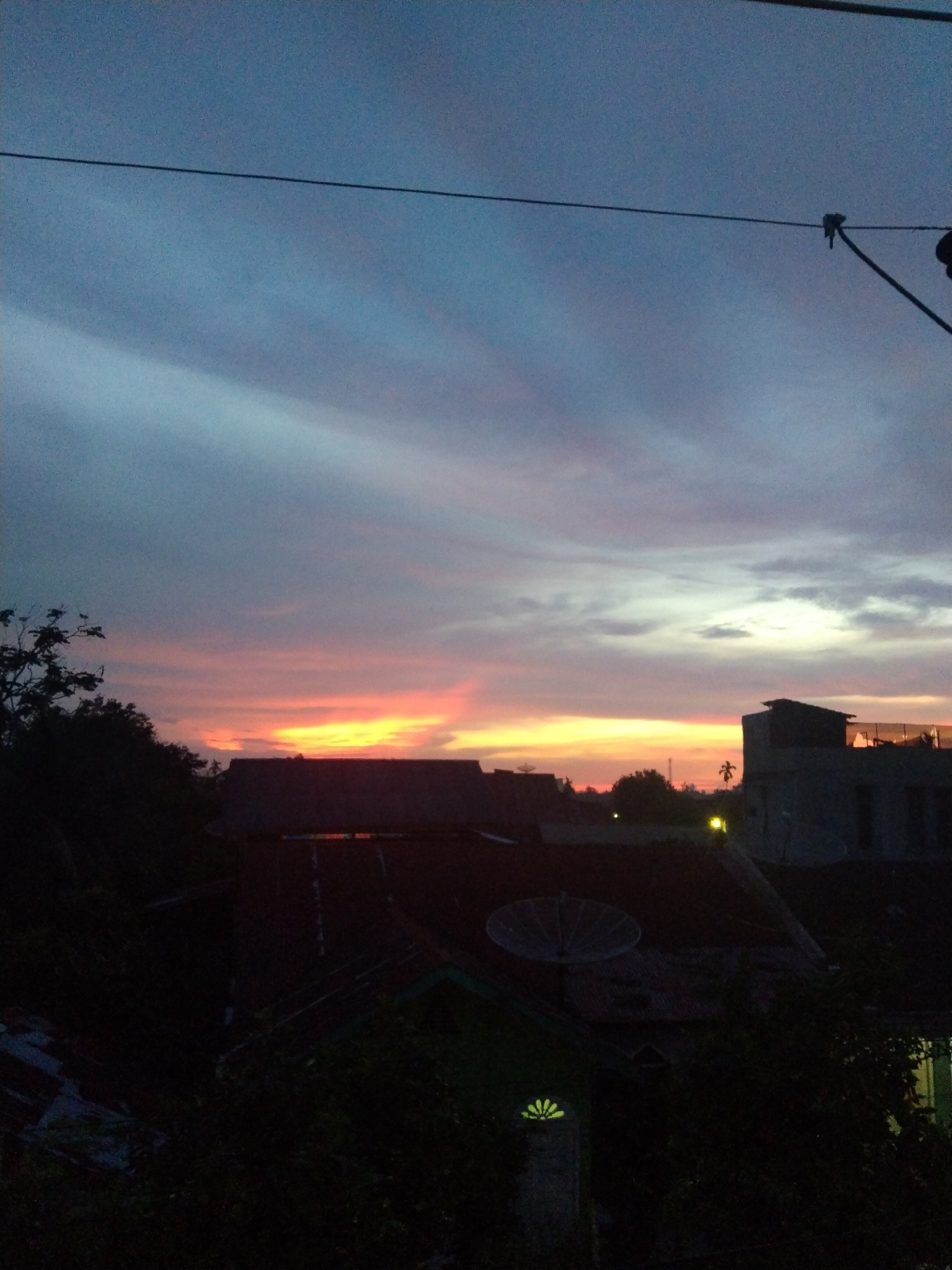 The image shows a sunset sky over the city of Bireuenmeunasahdayah, Indonesia. The sky exhibits shades of blue, gray, orange, and red. In the foreground, there are silhouettes of rooftops and trees. One rooftop features a satellite dish. Power lines run across the top of the frame. A few lights are visible amongst the buildings.