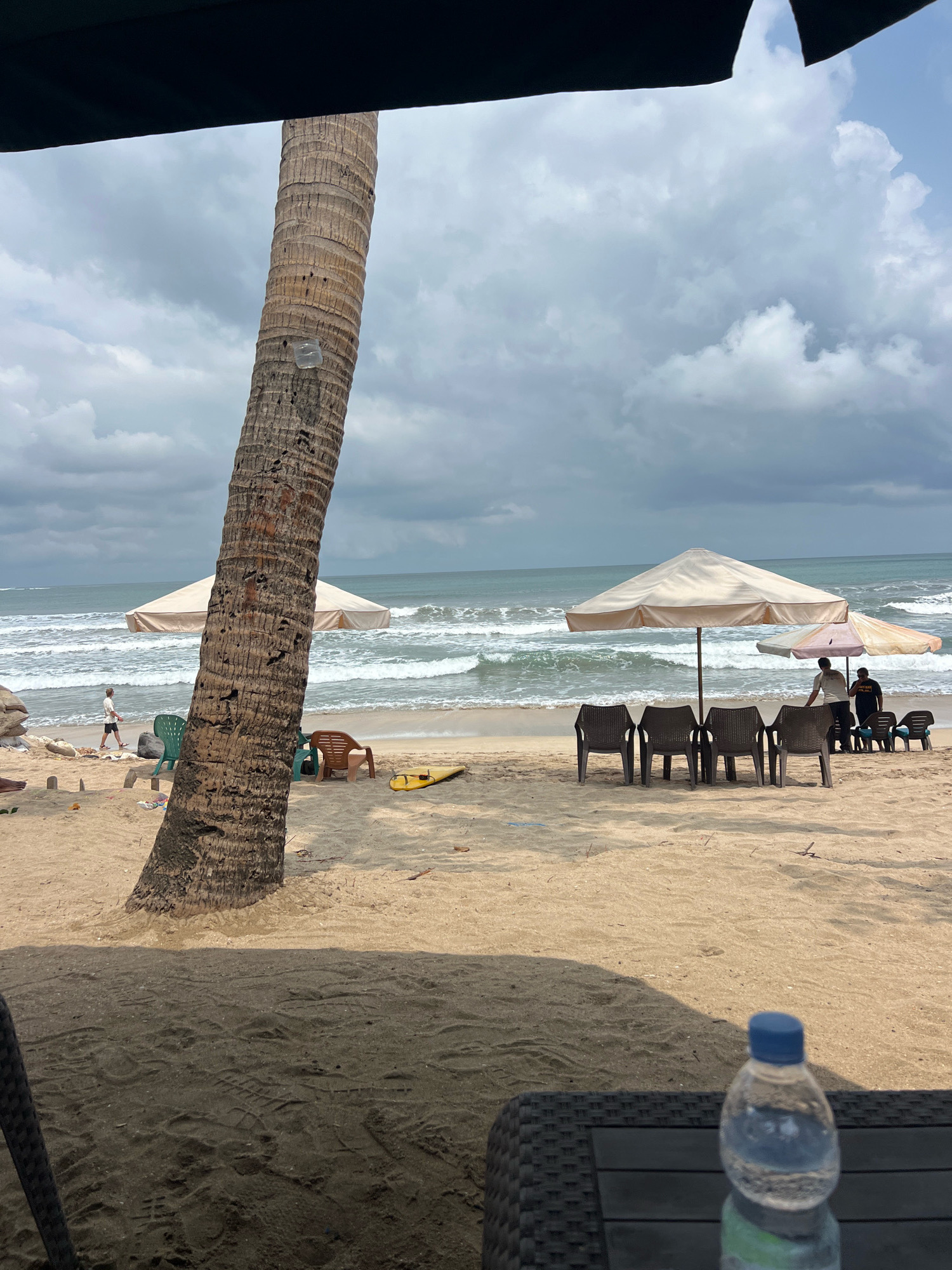 A man walks on a sandy beach in Kuta, Indonesia, with palm trees and umbrellas in the foreground and the ocean in the background. The sky is cloudy. There are chairs set up on the beach.