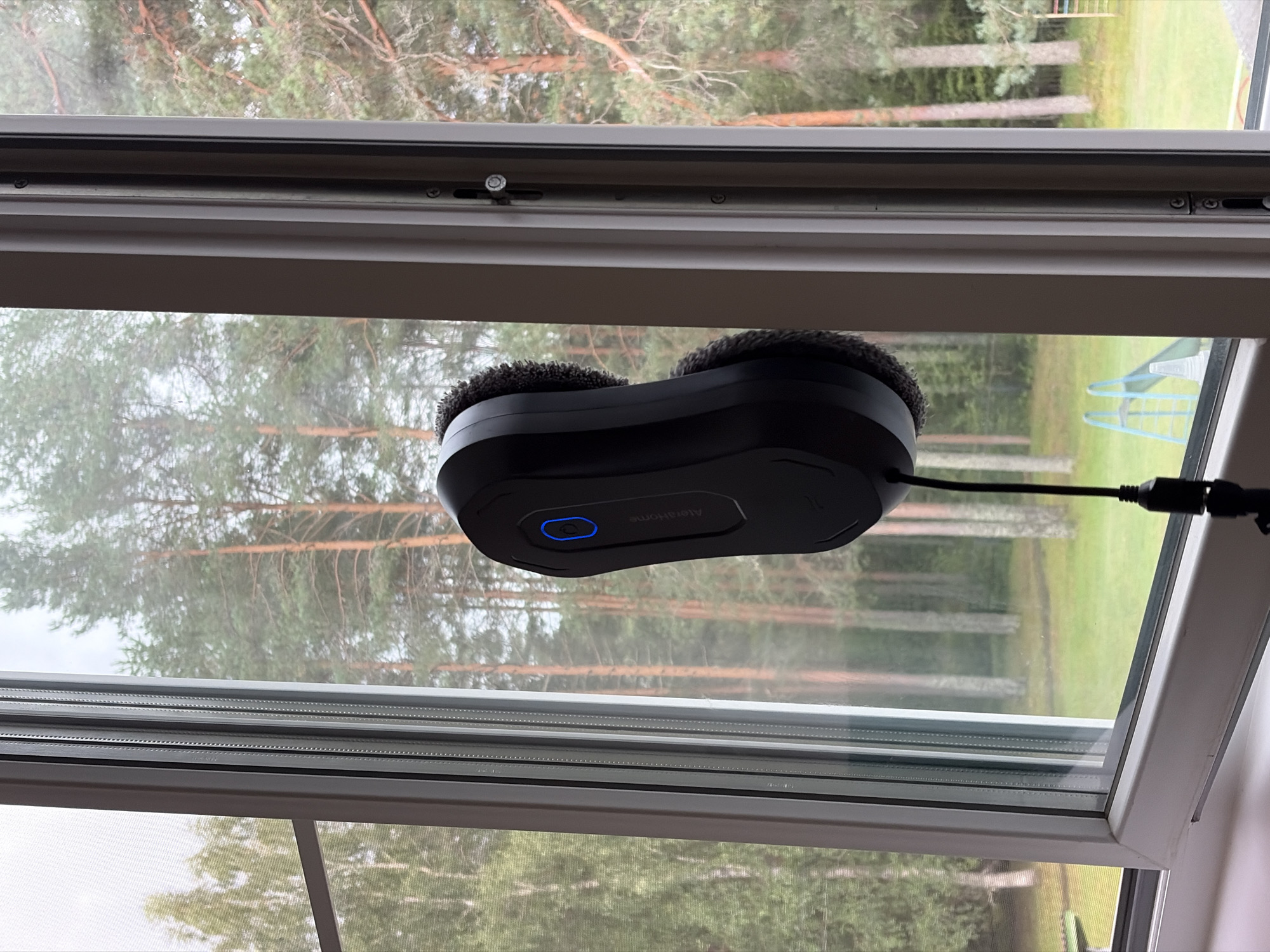 A black robot window cleaner is attached to the glass of a window, positioned vertically. The cleaner has two rotating pads, one visible on the left and another on the right, which are fuzzy and grey. A blue illuminated power button is present on the front of the device. A black cord extends downwards from the cleaner, connected to a power adapter. The window offers a view of a wooded area with tall pine trees. In the background, a green lawn and a children's play structure with blue slides and a white platform are visible. The scene appears to be a residential setting, possibly in Ганьково, Russia, given the context. The overall lighting suggests an overcast day.