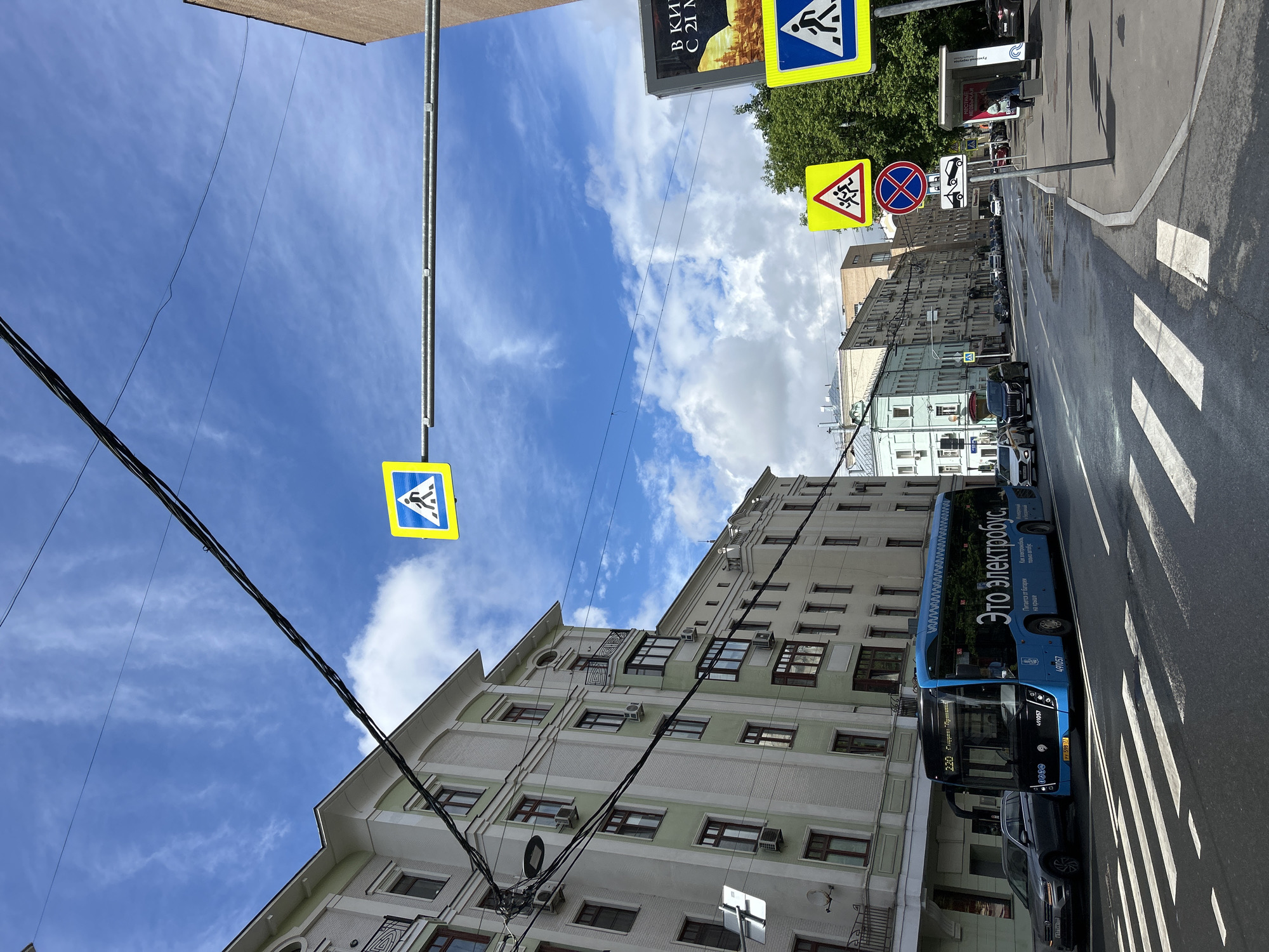The image shows a street scene in what appears to be a city, possibly in Russia, given the Cyrillic text on the bus. The scene is oriented vertically. In the foreground, there is a pedestrian crosswalk with white stripes on the pavement. A blue bus is parked adjacent to the crosswalk, bearing the inscription "Это электробус," indicating it is an electric bus. The side of the bus also has other Russian text and a vehicle identification number (491057). A dark-colored car is parked partially obscured in front of the bus.
In the background, a light-colored building with multiple windows and architectural details occupies a significant portion of the frame. Above the street, there are black power lines running across the sky. A blue and yellow pedestrian crossing sign hangs from a pole. Further down the street, more traffic and buildings can be seen.
The sky is visible above the buildings and is partially cloudy with a mix of blue and white. Several street signs are posted on the side of the road indicating crosswalks. The image appears to be taken during daylight under fair weather conditions. The location is urban and relatively dense.