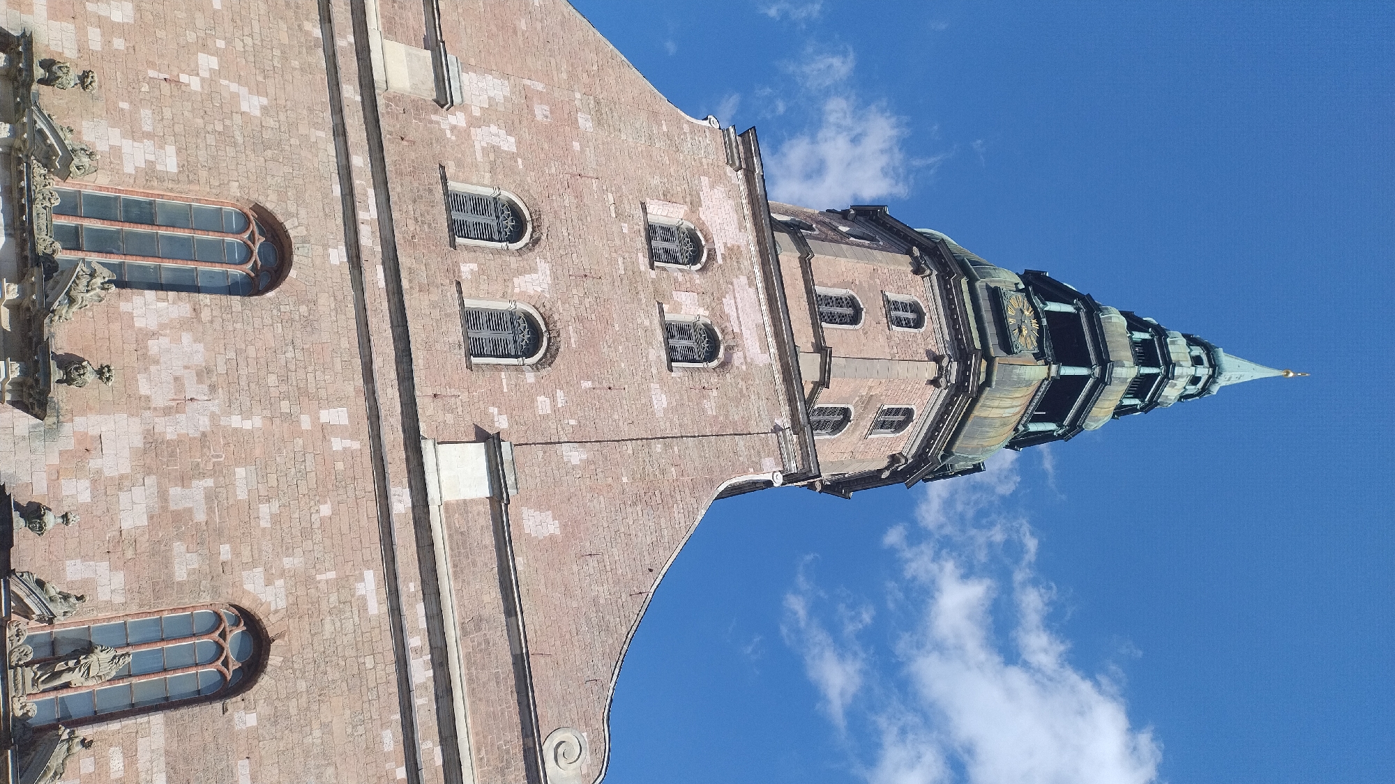 The image shows a low-angle, close-up view of the facade and steeple of St. Peter's Church in Riga, Latvia. The church is constructed of pinkish-red brick, with arched windows featuring dark shutters. The steeple is multi-tiered and topped with a pointed spire, adorned with copper elements that show signs of weathering. The sky above is a bright blue, with scattered white clouds. The architectural style is Gothic, characteristic of many historic European churches. There are no people visible in the image.