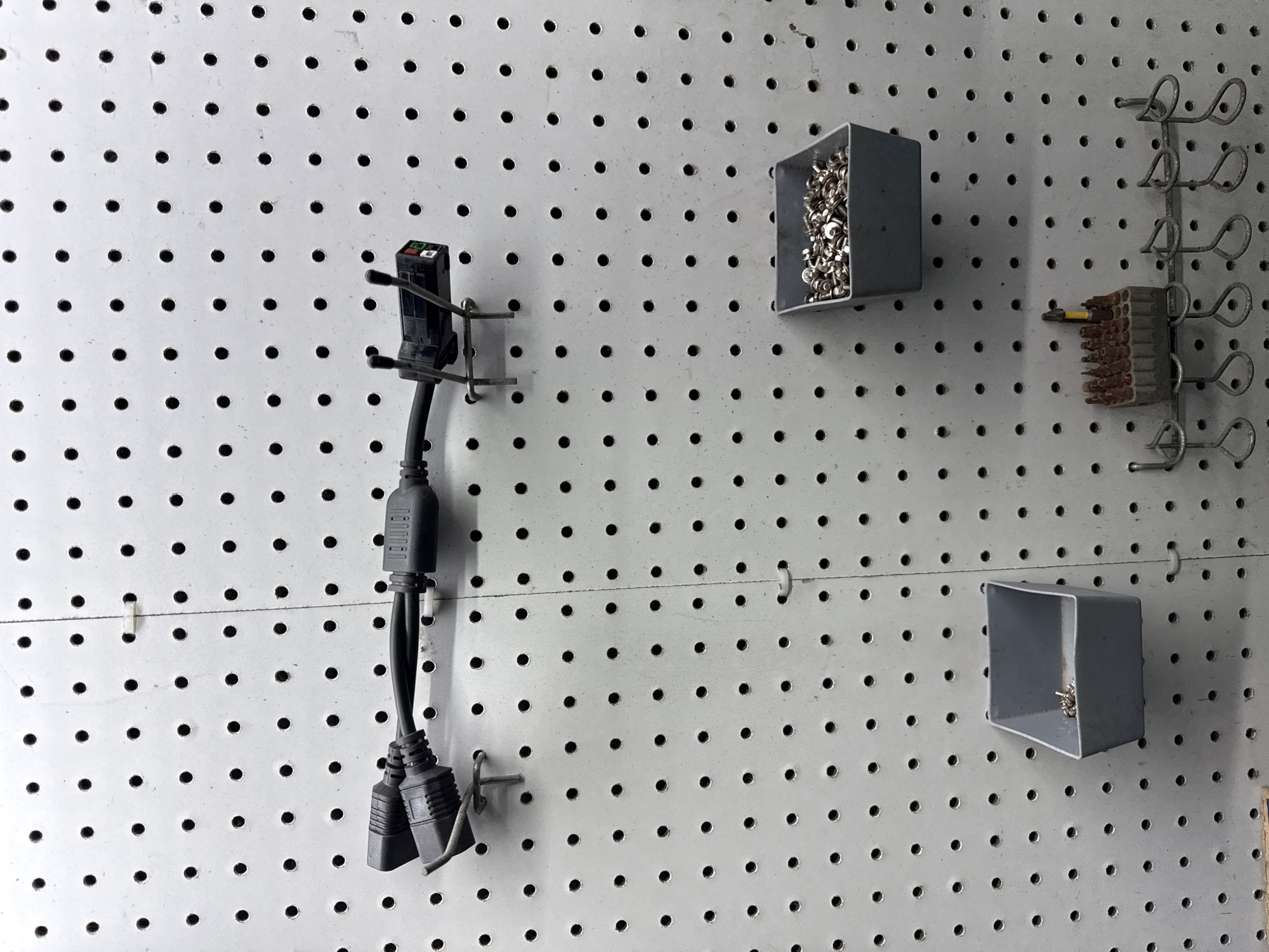 This image depicts a pegboard wall in what appears to be a workshop or storage area, likely located in Indiantown, United States of America. The pegboard is white and features a uniform pattern of circular holes.
Mounted on the pegboard in the upper section are two black electrical cables connected to a power strip or adapter. The cables are secured to the wall with metal hooks. Below the cables, to the right, is a grey plastic bin filled with small metal fasteners, possibly screws or bolts.
Further down on the pegboard, to the left, is another grey plastic bin, this one containing a smaller quantity of similar metal fasteners. Below this bin is a rack made of wire that holds a set of drill bits or screwdriver bits in a grey plastic holder. Several other wire loops, possibly for hanging tools, are also visible on this rack.
The overall scene is one of organized storage for electrical components and hardware. There are no people visible in the image.