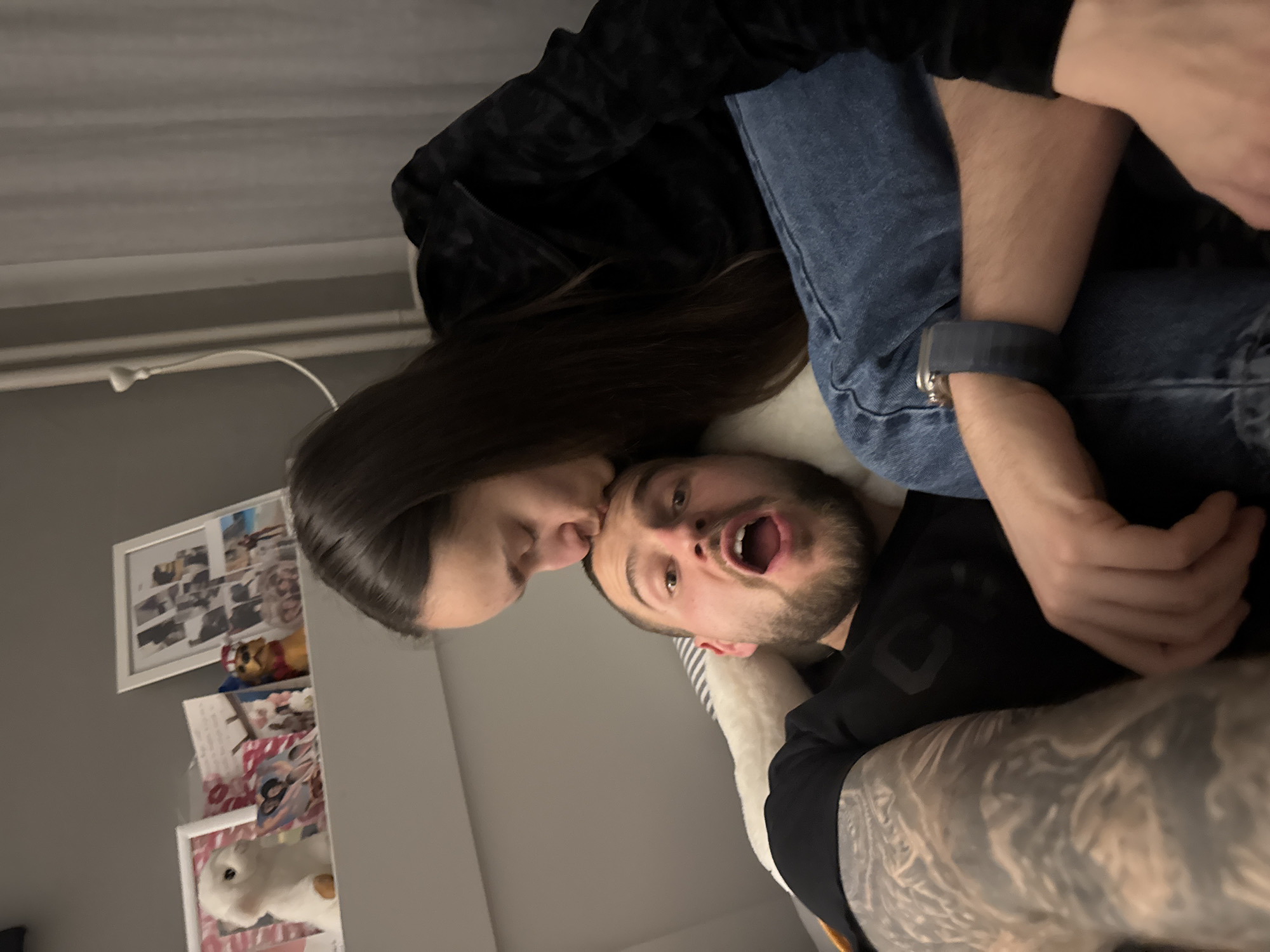 A woman is kissing a man on the forehead. The man has a beard and his mouth is open. He is wearing a black shirt. The woman has long, dark hair and is wearing a black hooded jacket and blue jeans. The man is wearing a smartwatch. There is a white stuffed animal and some framed photos on a shelf in the background. The wall is grey. The image appears to be taken indoors, perhaps in a home. It is not possible to confirm the location as Sofia, Bulgaria.