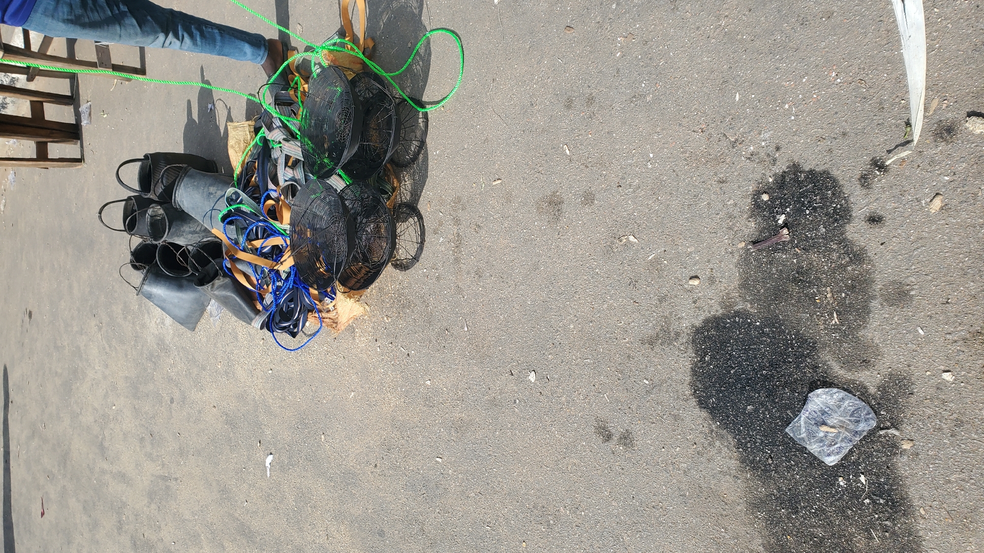The image depicts an outdoor scene on an asphalt surface, likely in Jalingo, Nigeria, characterized by scattered items and a person partially visible. In the upper right, a person wearing blue jeans and brown footwear stands, holding a green rope. In the central area, a collection of black plastic buckets with handles are piled together. Adjacent to the buckets is a bundle of ropes and straps, including blue and brown cords, intertwined with what appears to be a beige fabric or mat. Below this collection, two circular, black wire baskets are stacked on top of each other, with a third basket slightly offset. The ground shows evidence of wear and tear, with patches of dark staining and small debris, including a piece of clear plastic packaging containing an unknown object and a small bone-like fragment, visible in the lower portion of the frame. A wooden structure is partially visible in the upper right background. The lighting suggests it is daytime, with shadows cast on the ground.