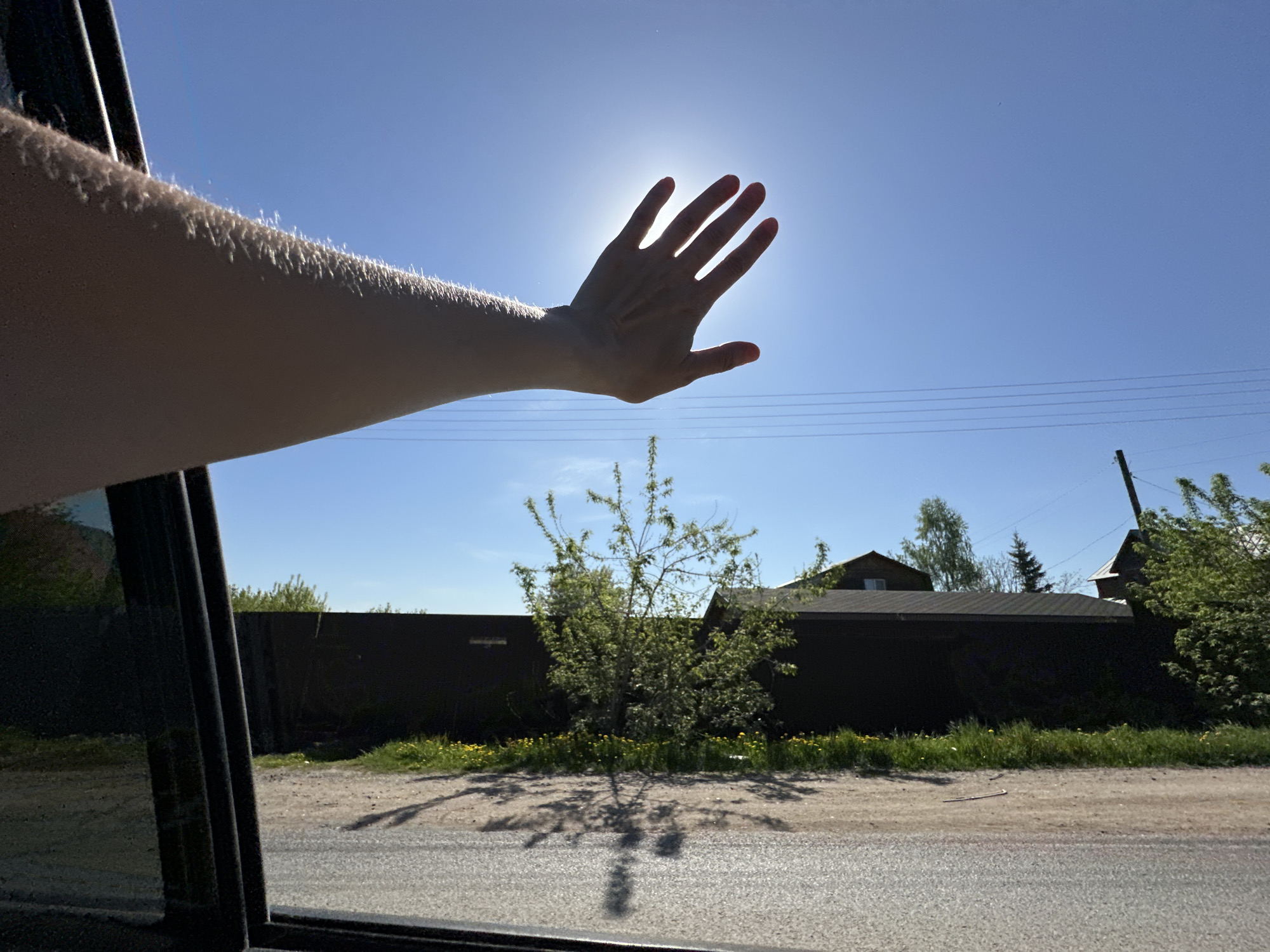 An arm extends from the open window of a vehicle, the hand outstretched towards the sun. The scene viewed through the window includes a low, dark fence, a small tree, grass with yellow flowers, a gravel road, and houses in the background. Electrical lines run across the clear blue sky.
