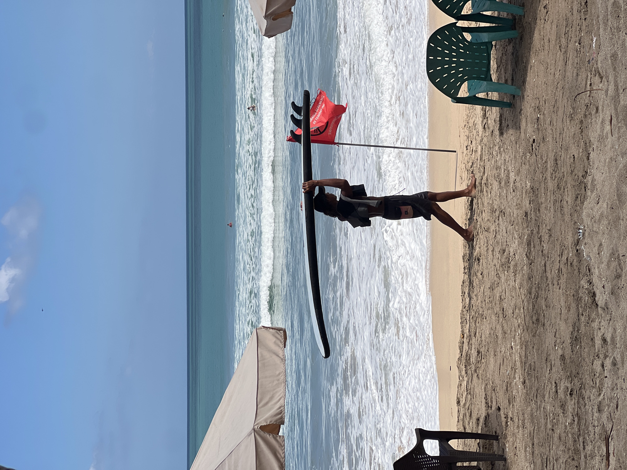 A man is walking on the beach of Kuta, Bali, Indonesia, carrying a black surfboard on his shoulders. He is wearing dark shorts and a dark shirt draped over his shoulders. Beside him, a red flag with white writing and three black fins is planted in the sand, indicating a dangerous area for swimming. In the background, the turquoise ocean with white waves stretches to the horizon under a clear blue sky with a few scattered clouds. Several people are visible in the water, some appearing to be surfing. Umbrellas and plastic chairs are set up on the sandy beach, suggesting a tourist area.