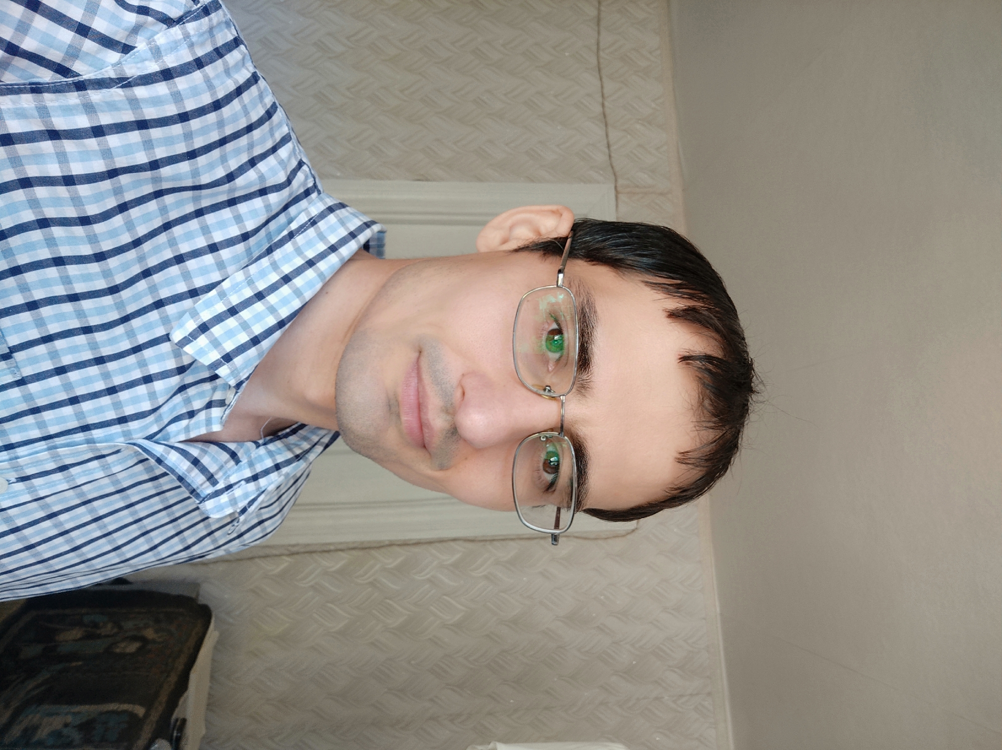 A close-up, head-and-shoulders portrait shows a man wearing glasses and a blue and white checkered collared shirt. He has dark hair and a fair complexion. The background is a wall with a textured pattern. The lighting suggests an indoor setting, possibly during the day.