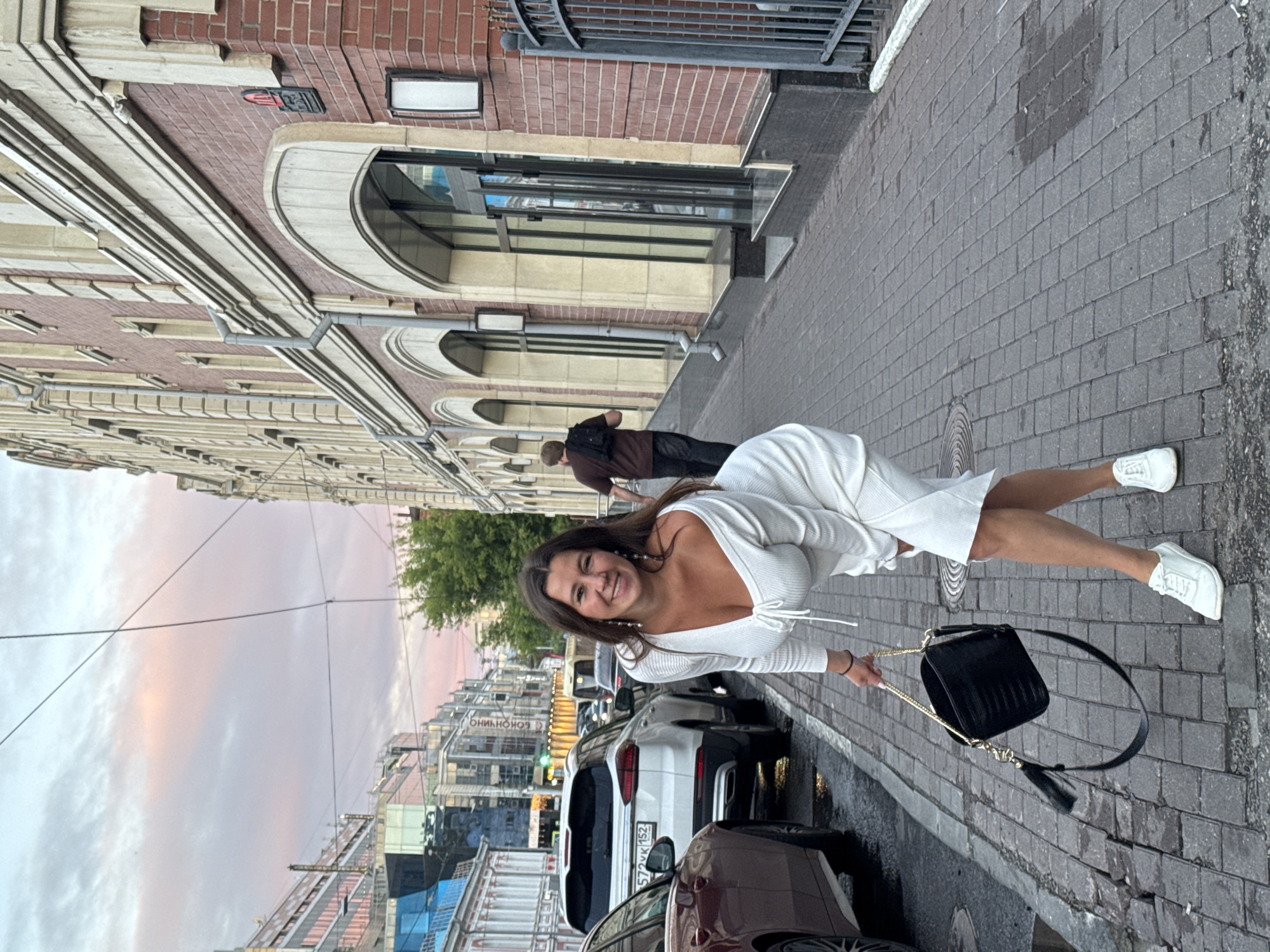 The image depicts a woman posing on a cobblestone sidewalk in front of a brick building with arched windows. She is wearing a white, off-the-shoulder dress and white sneakers, and she is holding a black handbag. Cars are parked along the street to her left, and a man is walking away in the background. The sky is a soft pink and orange, suggesting it is sunset or sunrise. The building appears to be an older European-style structure with decorative architectural elements. The overall impression is of a street scene in an urban environment. The building visible in the background with the text "БОНЧИНО" strongly suggests the location is in Nizhny Novgorod, Russia, specifically on Bolshaya Pokrovskaya Street.