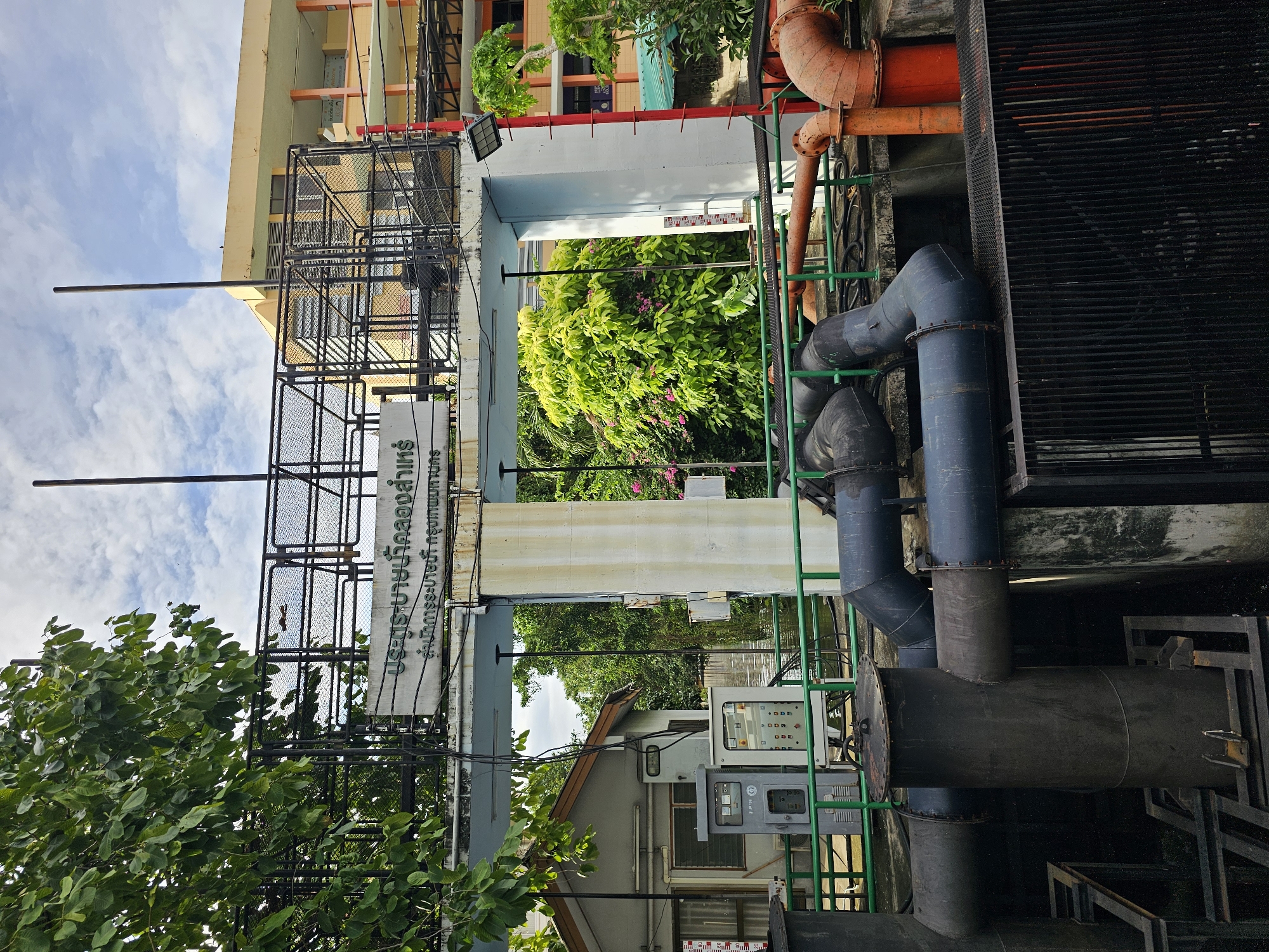 A medium shot captures an outdoor scene at a water management facility in Bangkok, Thailand. The focal point is a large, light-colored concrete structure that appears to be part of a pumping station or sluice gate system. Overhead, a dark metal frame structure with mesh infill spans across, supporting a white sign with Thai text that reads "ประตูระบายน้ำคลองลาม" which translates to "Lam Canal Sluice Gate." Below the sign, smaller Thai text reads "สำนักการระบายน้ำ กรุงเทพมหานคร" which translates to "Department of Drainage and Sewerage, Bangkok Metropolitan Administration."
On the left, a small, grey building with a peaked roof is visible, housing electrical control panels and equipment. A green metal railing runs along the foreground, separating the viewer from the pumping equipment. Several large, dark grey pipes, some bent in curved sections, are prominent, leading into and out of the concrete structure. To the right, a bright orange pipe section is visible, suggesting different water flow systems.
Behind the concrete structure, lush green trees and foliage dominate the background, with a hint of a light yellow building visible in the distance through the branches. The sky above is partly cloudy. The scene is well-lit, suggesting it is daytime. There are no people visible in the frame.