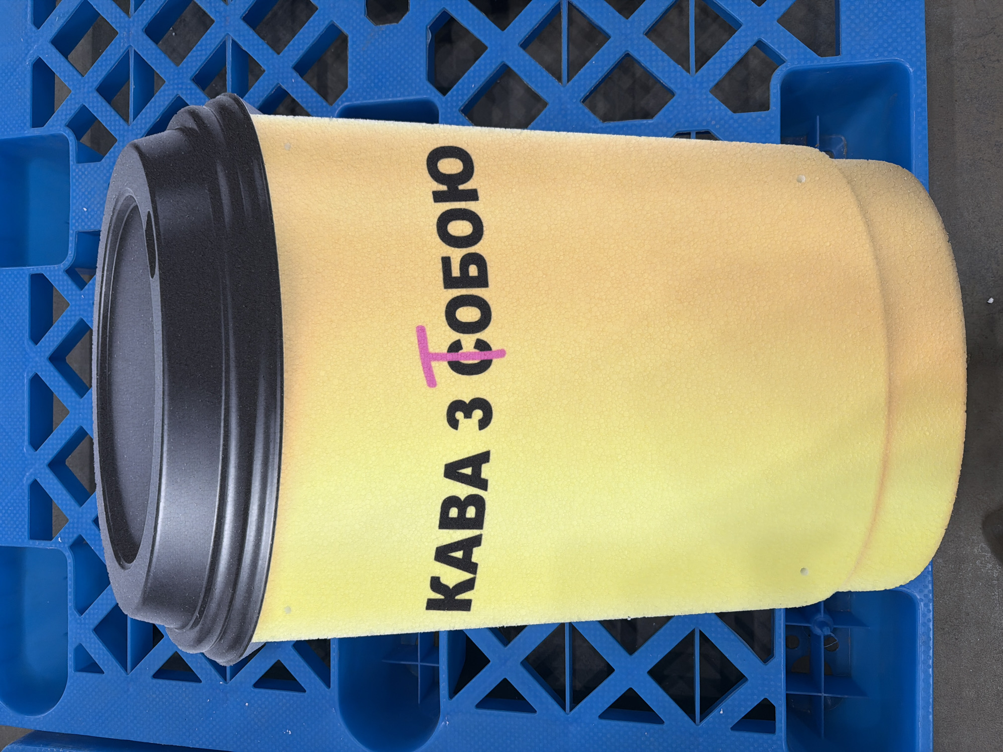 A close-up, eye-level shot shows a yellow foam coffee cup with a black lid. The cup has black Cyrillic text that reads "КАВА З СОБОЮ," which translates to "Coffee to go." A pinkish mark is visible over the letter "T." The cup is positioned in front of a bright blue plastic crate with a diamond-shaped grid pattern. The background is dark and out of focus, suggesting an indoor or shaded outdoor setting. The image does not provide any specific location details within the city of Ternopil, Ukraine, only the presence of a coffee cup advertising "Coffee to go."