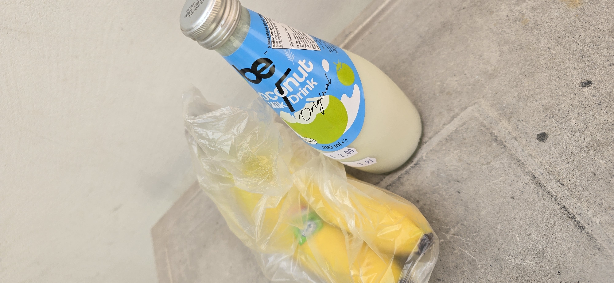 The image shows a bottle of "be Coconut Milk Drink Original" and a plastic bag containing bananas. The bottle has a silver cap and a label with blue and green text. The bananas are yellow and appear to be slightly bruised. Both items are resting on a concrete surface, possibly a step or sidewalk. The exact location in Burgas, Bulgaria, cannot be determined from the image alone. There are no people visible.