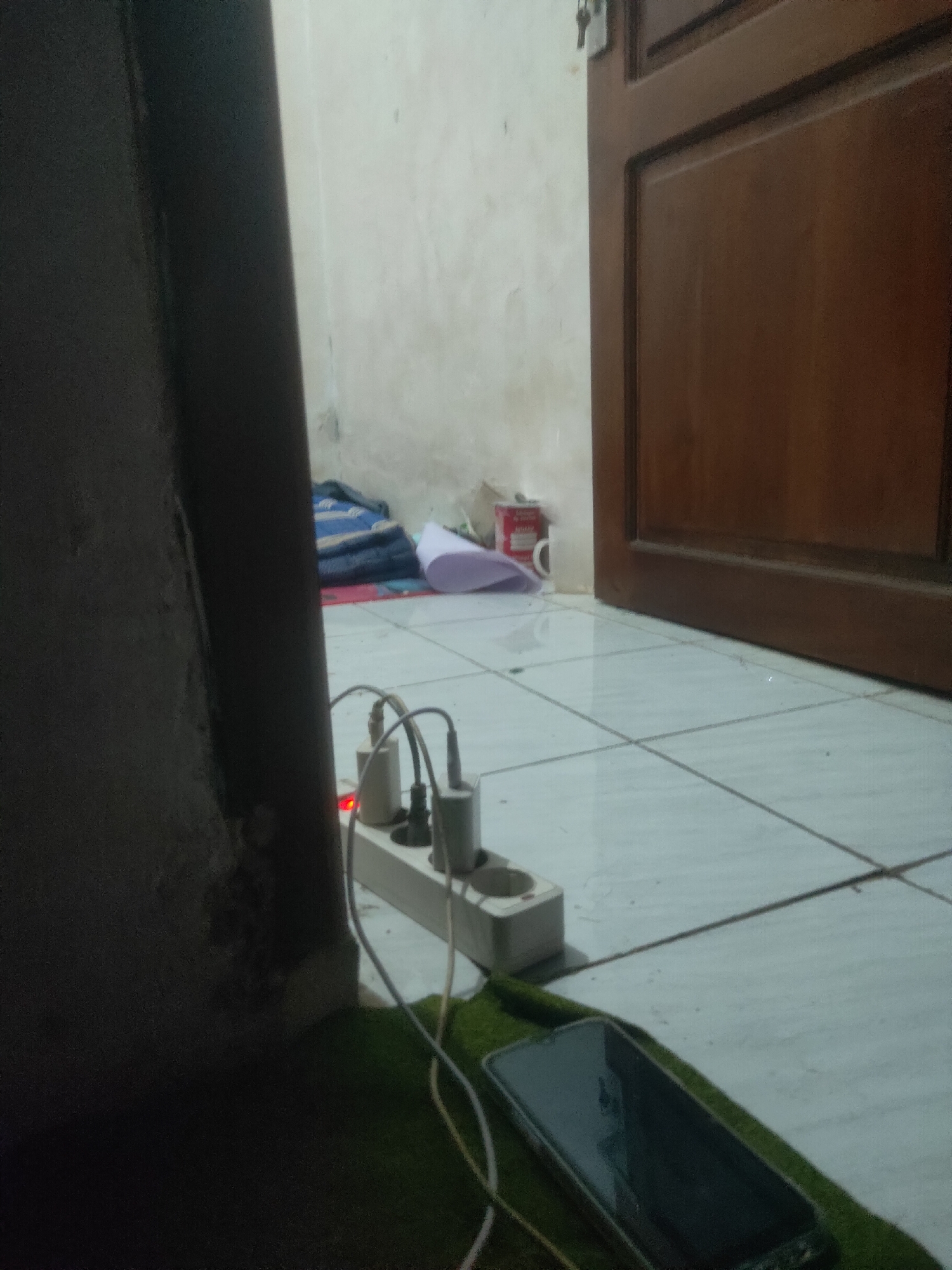 The image shows a close-up of a power strip with three phone chargers plugged in, two of which are connected to a smartphone lying on a green mat. The power strip is plugged into a wall socket on a tiled floor. In the background, there is a wooden door and a wall. The scene appears to be an indoor setting in a home or lodging, likely in Antirogo, Indonesia, given the context. There are no people visible in the image.