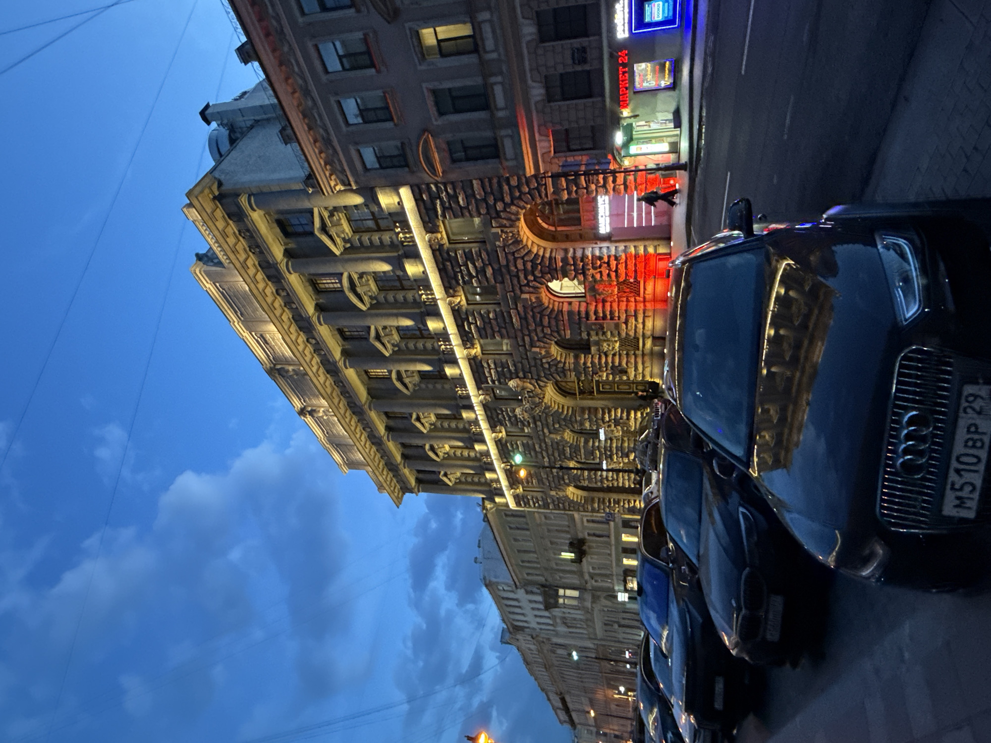 This image depicts an urban street scene at dusk or night, set in Saint Petersburg, Russia. The sky is a deep blue with scattered clouds, indicating either late evening or early morning twilight.
On the left, a large, ornate multi-story building with classical architectural elements dominates the view. It features prominent columns, pilasters, and decorative cornices, illuminated by warm artificial light that accentuates its intricate details. The lower part of this building has a rustic stone facade with arched windows and entrances. One such entrance is strikingly lit with red neon lights, revealing what appears to be a commercial establishment. A solitary person is visible near this red-lit entrance.
Along the right side of the street, a line of cars is parked. The closest vehicle is a dark Audi SUV, whose front grille and headlights are clearly visible. Its Russian license plate, "M510BP 29", confirms the country context. Reflections of the illuminated architecture shimmer on the vehicle's dark surface. Other cars are visible extending further down the street.
In the background, another building with a more modest facade can be seen. Streetlights cast soft glows, and numerous overhead power lines or tram lines crisscross the blue sky.
Visible text includes "МАРКЕТ 24" (Market 24) and "DOPO" on various neon signs. Another sign above an arched entrance on the main building appears to read "MANCHINYC" or "МАНЧИНЫ". The scene conveys the calm yet active atmosphere of a city street during the evening hours.