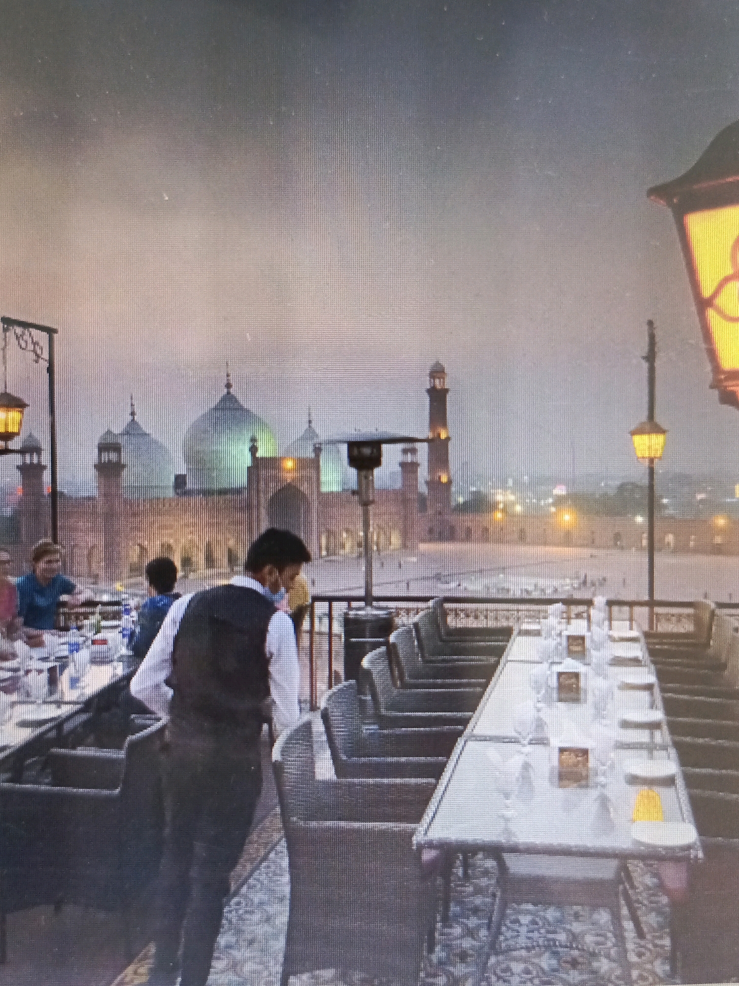 This image captures a rooftop dining scene overlooking the Badshahi Mosque in Lahore, Pakistan. The foreground shows a long table set with white plates, glasses, and small table tents. Several chairs with dark upholstery surround the table. A man in a white shirt and dark vest, presumably a waiter, stands near the table, facing away from the camera. To the left, other diners are seated at tables, with one woman in a blue top visible. In the background, the imposing Badshahi Mosque dominates the skyline with its distinctive domes and minarets, illuminated by evening lights. The sky is overcast, contributing to a muted, atmospheric light. The overall scene depicts a serene evening setting for dining with a prominent historical landmark as the backdrop.