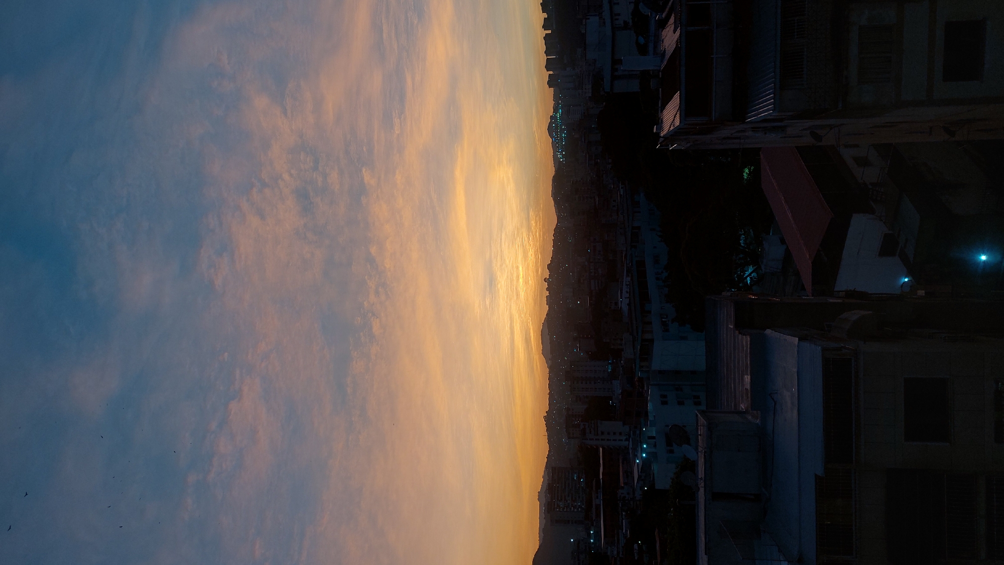 The image shows a sunset view from a building in Caracas, Venezuela. The sky is predominantly blue with orange hues as the sun sets. Below the horizon line are hills and densely populated areas with buildings lit by streetlights. The right side of the image depicts a residential neighborhood with rooftops and building structures, where buildings get darker with less light. Birds are visible in the sky.