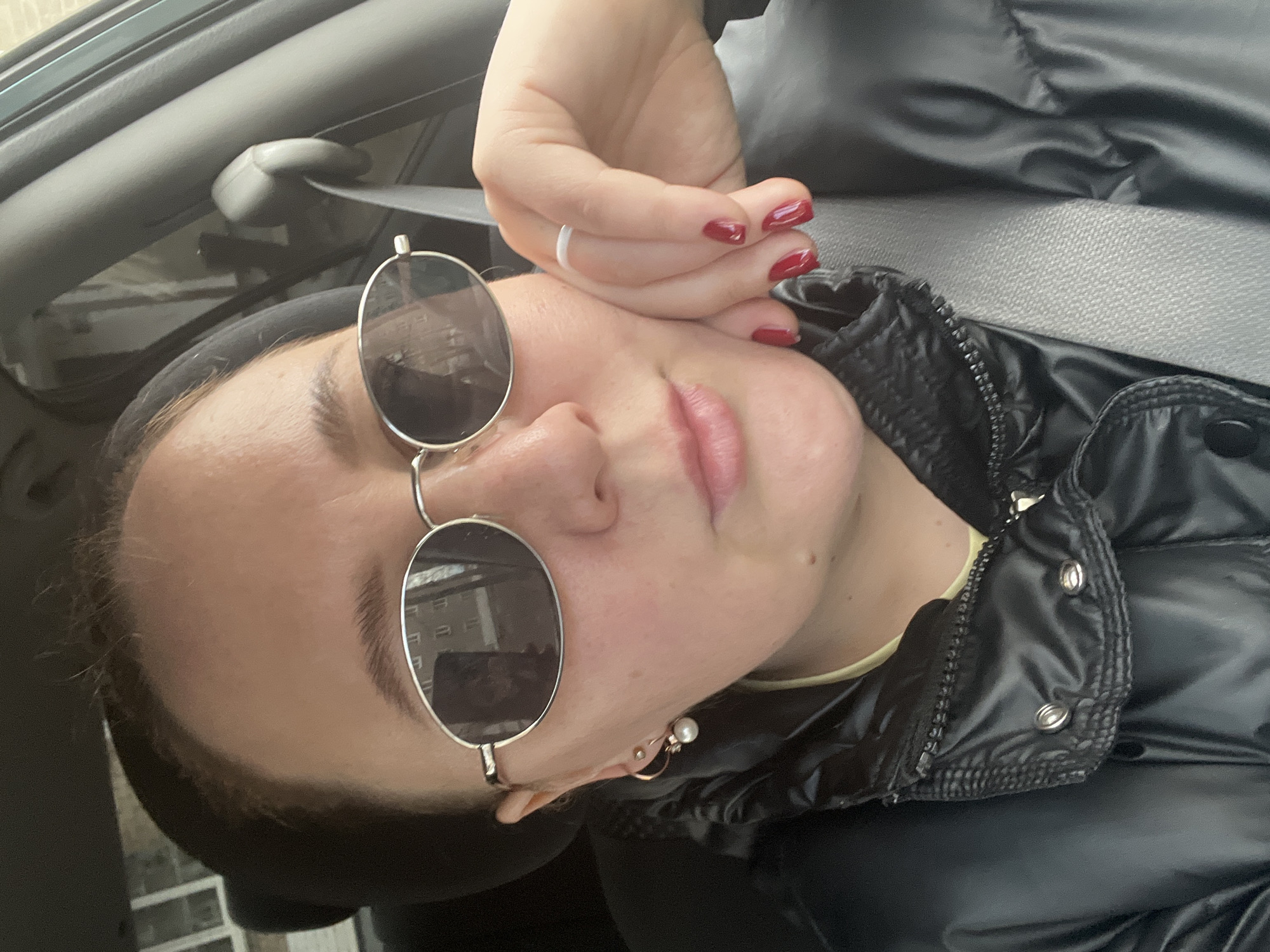 A person, likely female, is depicted in a close-up, slightly tilted portrait, resting inside what appears to be a car in Chaikovsky, Russia. The individual wears round, silver-framed sunglasses, partially obscuring the upper face. Her mouth is slightly open, and a pearl earring is visible on her right ear. Her right hand, adorned with glossy red nail polish and a plain silver ring on the ring finger, rests against her cheek. She is dressed in a black, shiny puffer jacket. Portions of a car seatbelt and the car's interior roof are visible in the background, reinforcing the automotive setting. The sunglasses reflect what seem to be building windows or architectural elements. The expression conveys a relaxed or pensive mood. The lighting is soft, not indicating a specific time of day or weather. No discernible text is present in the image.