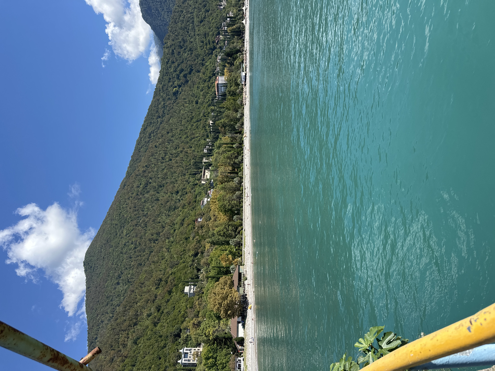 The image shows a panoramic view of a beach and the sea in Abaata, Abkhazia. The turquoise water fills the foreground, with gentle ripples on its surface. A white pebble beach lines the shore, with scattered sunbathers and a few people wading in the water. Behind the beach, a line of trees and buildings runs along the base of a lush, green mountain. The mountain is densely covered with trees and rises steeply towards a clear blue sky dotted with fluffy white clouds. In the lower left corner, a yellow railing and a green leafy plant are visible, suggesting the photo was taken from a pier or balcony overlooking the scene. Several houses and hotels are visible nestled on the hillside, with some architecture suggesting a resort town. The overall scene depicts a sunny day at a coastal resort.