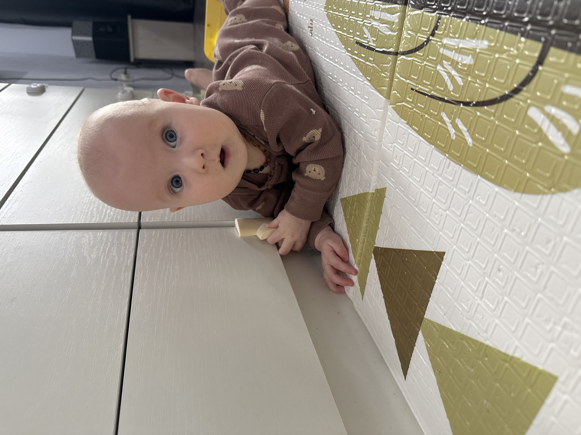 A baby in a brown patterned onesie with a beaded necklace lies on a white surface with a yellow foam mat to their right, which has black and yellow animal figures. The baby has blue eyes and a surprised expression, holding a small white object. The scene appears to be indoors, with furniture visible in the background. There is no indication of the location being in Yakutsk, Russia.