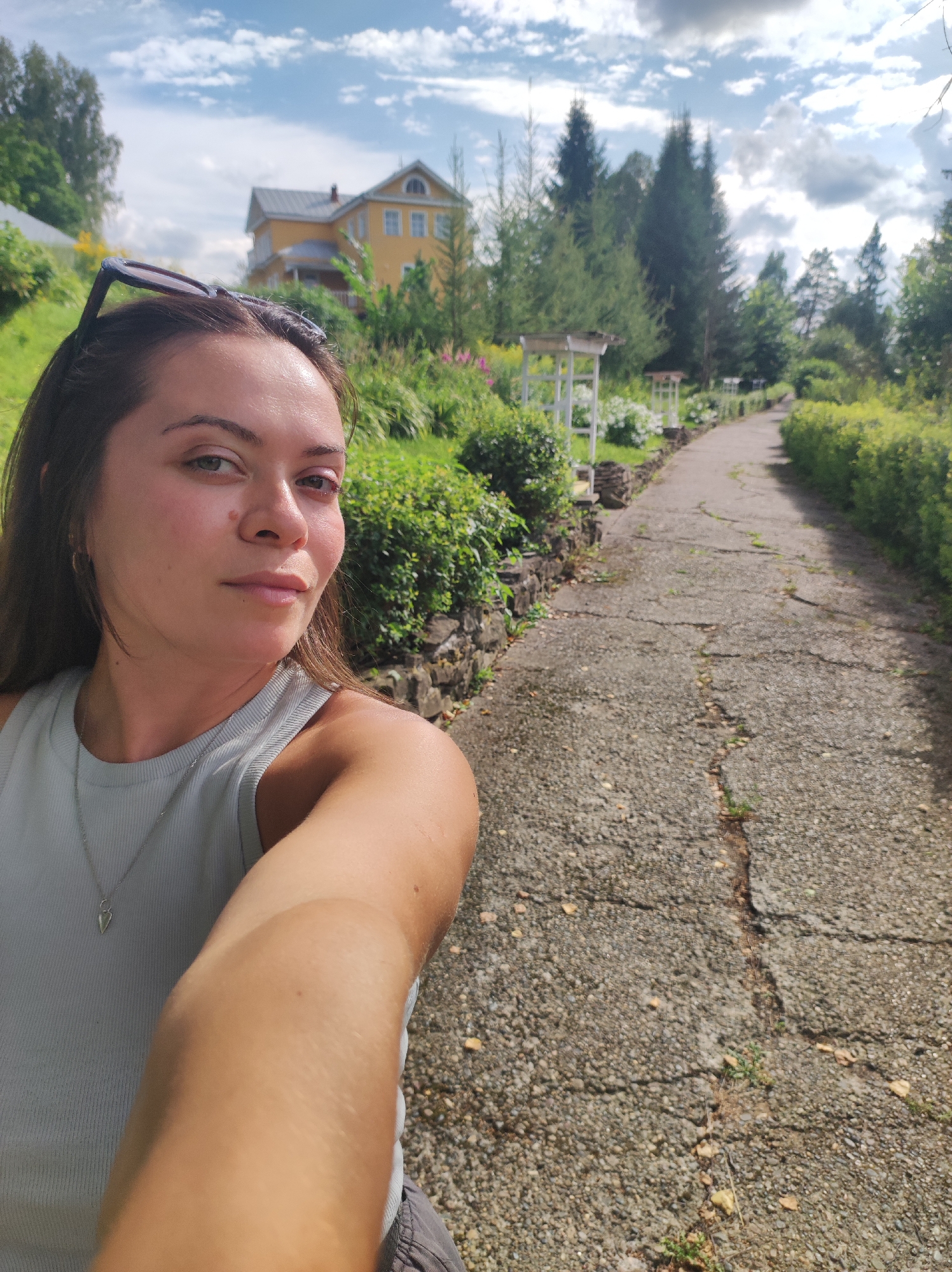 A woman is taking a selfie outdoors on a sunny day. She is wearing a light blue tank top and a silver necklace with a pendant. Sunglasses are perched on top of her head. Behind her, a paved path winds through a garden with lush green bushes and flowers. In the distance, a two-story yellow house with a grey roof is visible, surrounded by trees. The sky is blue with scattered white clouds. The image appears to have been taken in Zhigalki, Russia, given the architectural style of the house and the surrounding vegetation.