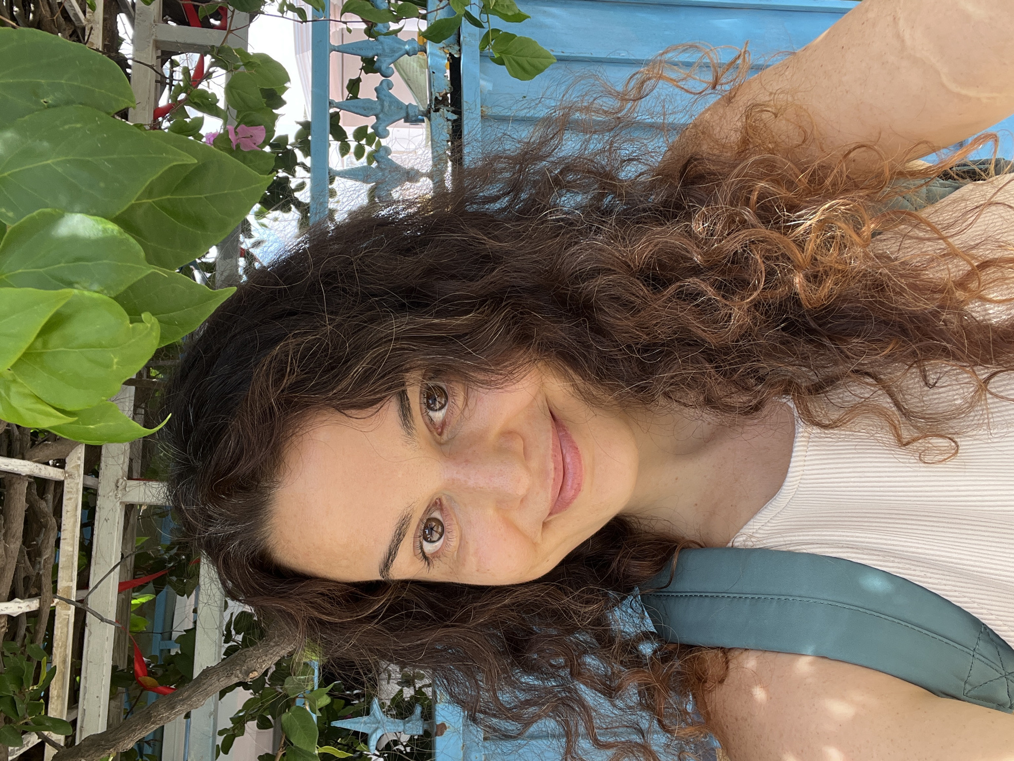 A medium shot shows a woman with curly brown hair smiling at the camera. She is wearing a light-colored ribbed tank top and a teal backpack strap is visible over her left shoulder. The background features a blue wooden fence and some greenery, including leaves and a pink flower. The scene suggests an outdoor setting, possibly in Mũi Né, Vietnam, known for its beaches and vibrant blue fishing boats, which could be implied by the blue fencing in the background.