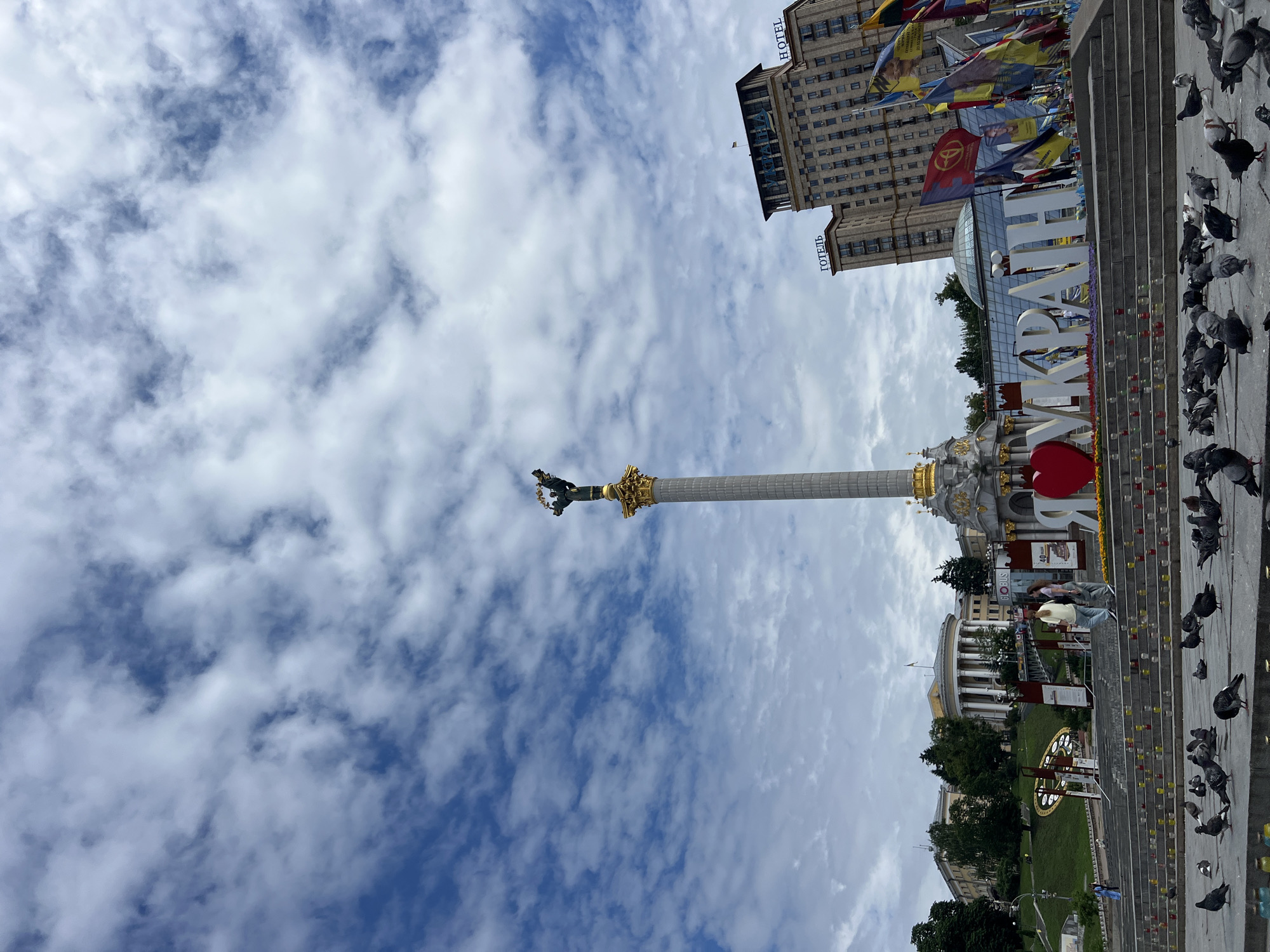 This is a full, eye-level shot of Maidan Nezalezhnosti (Independence Square) in Kyiv, Ukraine. The sky is mostly cloudy, with patches of blue visible. In the center of the frame, a tall, ornate monument stands, topped with a statue of a winged female figure holding a laurel wreath. In the foreground, a flock of pigeons are gathered on a stone plaza. Behind the monument, a large sign spells out "I LOVE UKRAINE" with a red heart at the beginning of the word "LOVE." Several flags are displayed behind the sign, including Ukrainian flags and other banners. In the background, the Hotel Ukraina building is visible, as well as other architectural structures and trees. The steps leading up to the monument are lined with candles, and two women are walking up the steps.