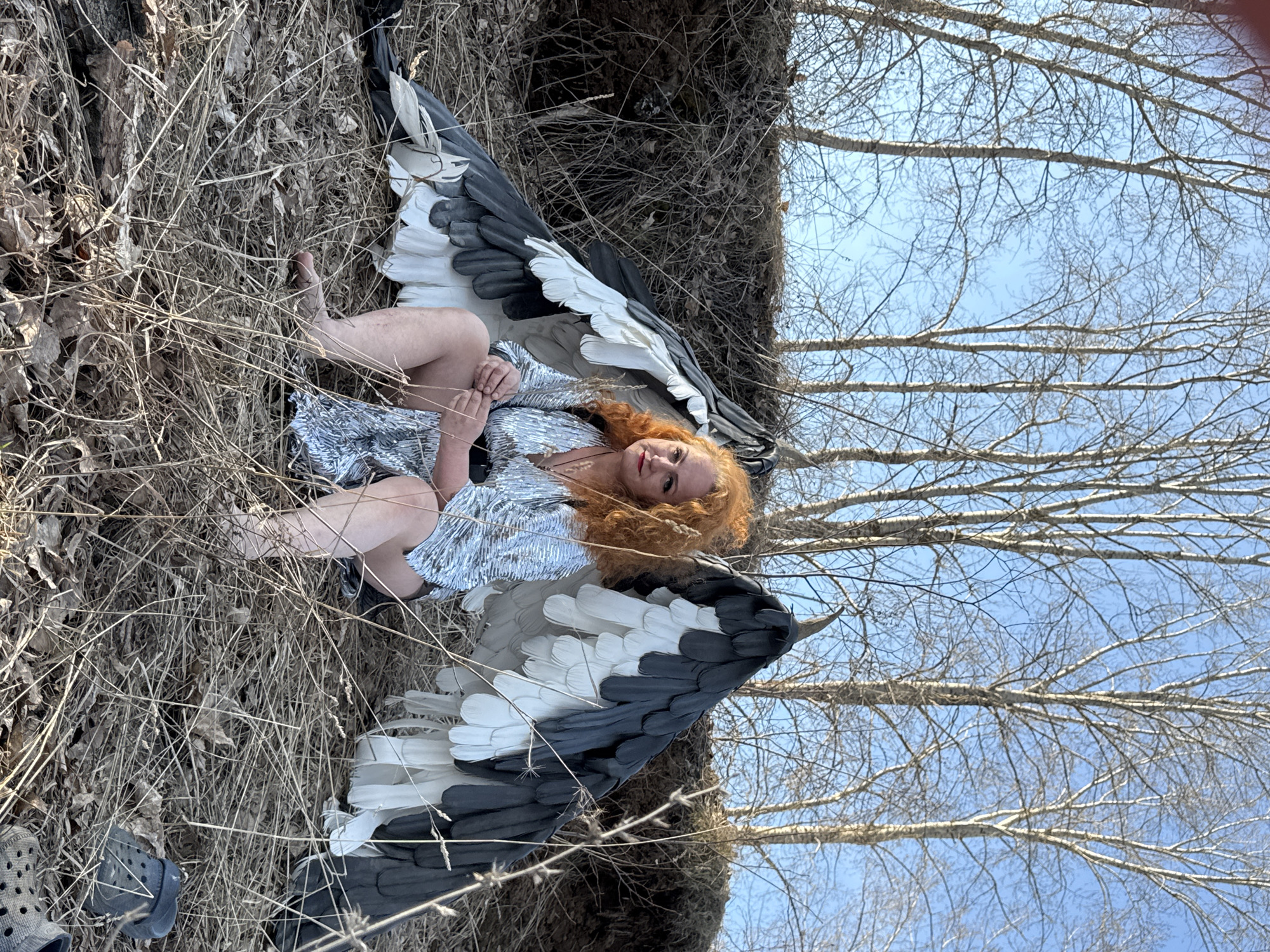 The image shows a woman lying on the ground in a natural setting, likely outdoors near a wooded area. She has red hair and is wearing a silver dress. The woman is adorned with large, decorative wings that are black and white. The ground is covered with dry grass and vegetation. In the background, there are bare trees against a pale blue sky. The image is oriented vertically, and the scene appears to be captured during daylight. The presence of the trees suggests the photo was taken near a park or wooded area. While the exact location within the city of Chaikovsky is not explicitly identifiable, the scene could be a location near the Kama river, as the Kama flows near Chaikovsky. Footwear, possibly Crocs, is seen in the lower left.