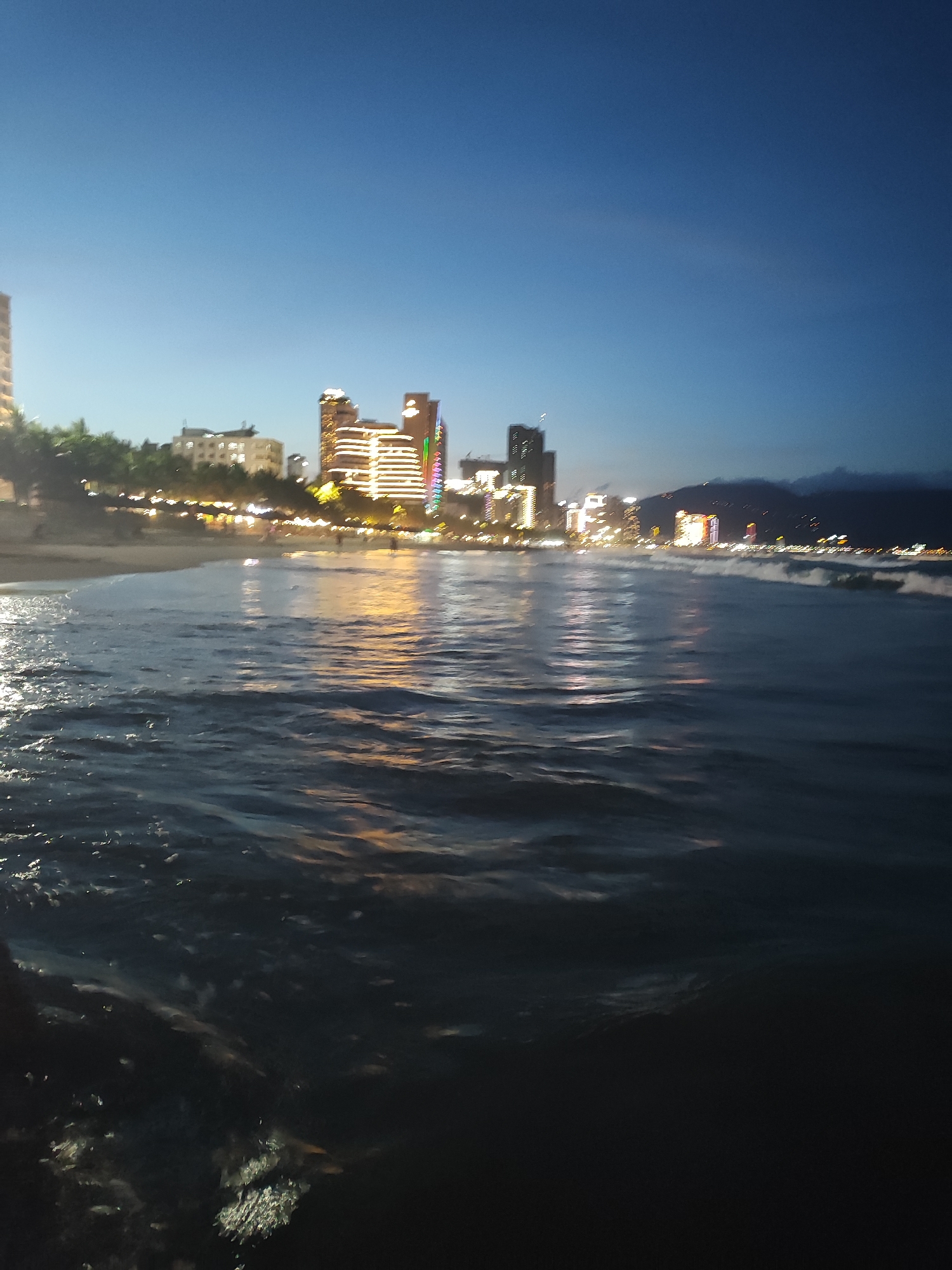 The image captures a nighttime view of the shoreline in Đà Nẵng, Vietnam. The foreground is dominated by the ocean, with visible waves reflecting the city lights. In the background, there is a blurred view of the city skyline, with illuminated buildings and a beach area. The sky is a gradient of blues, indicating twilight. The overall lighting conditions suggest it is dusk or a dark evening.