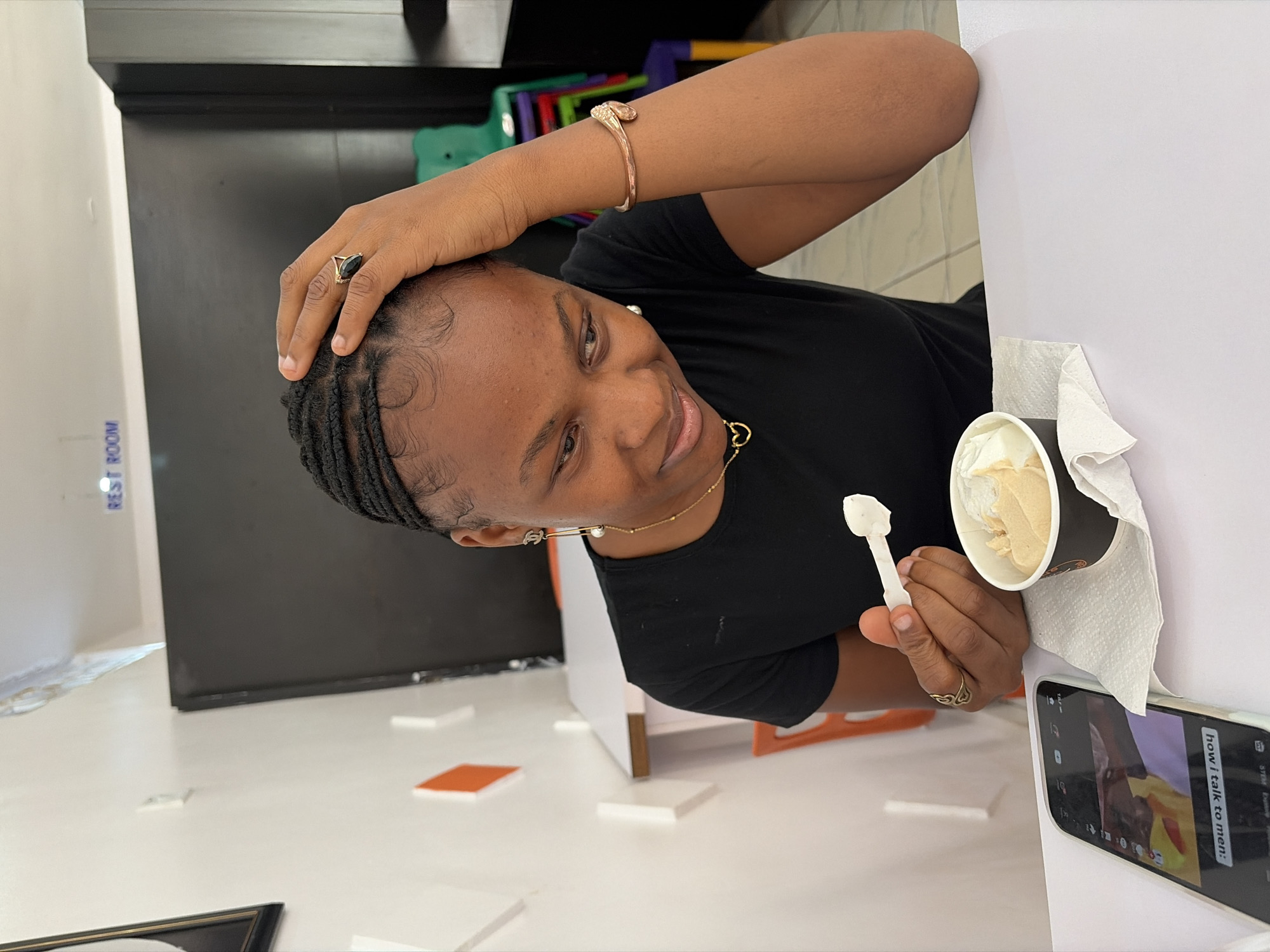 This image, taken at Bar Jirgi Summa, Nigeria, features a person, likely female, captured in a relaxed, indoor setting. The individual has dark skin and braided hair with styled edges, and is wearing a black t-shirt, a gold chain with a heart pendant, small stud earrings, a gold bracelet on the right wrist, and gold rings on both hands (one with a dark stone on the right index finger, another with a looped design on the left ring finger).
She is smiling and looks slightly off-camera, with her right hand gently touching her head. In her left hand, she holds a white plastic spoon, positioned over a black cup containing light-colored ice cream. A white paper napkin is tucked beneath the ice cream cup, which rests on a white surface next to a smartphone. The phone screen displays text that clearly reads "how I talk to men:," suggesting video or social media content.
The background includes a dark wall or structure behind the person. To the left, a white wall is visible with blue text partially reading "REST ROOM". The floor appears light-colored, possibly white, scattered with small, flat, square-shaped objects in white and orange. Further back, colorful, indistinct objects are visible, contributing to a casual and inviting atmosphere. The overall scene depicts a moment of leisure, enjoying a treat while likely browsing content on a phone.